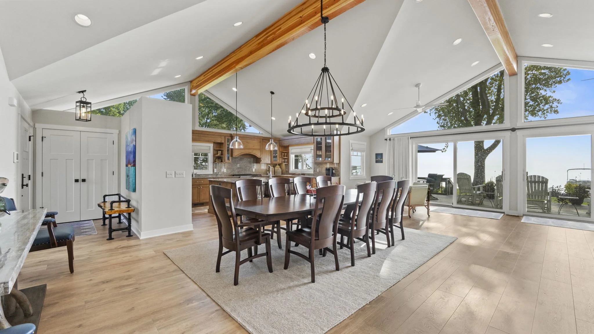 Open-concept dining and kitchen area with large windows, hardwood flooring, dining table with chairs, kitchen cabinets, and outdoor seating visible through sliding glass doors.