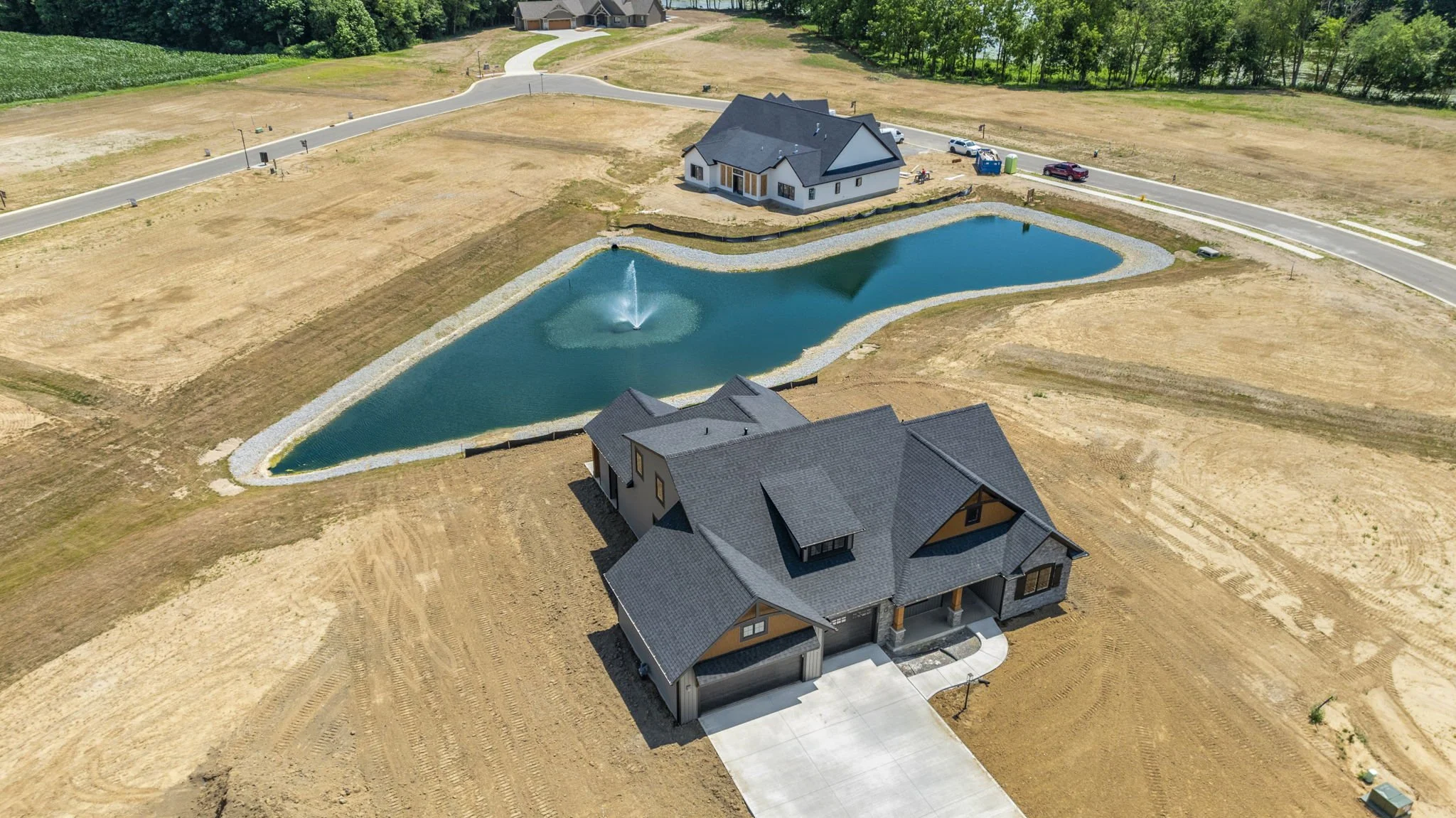 An aerial view of a new residential development showing two houses, a large pond with a fountain, and surrounding undeveloped land. One house is closer to the camera and has a driveway leading to a garage. The other house is farther back near the pon
