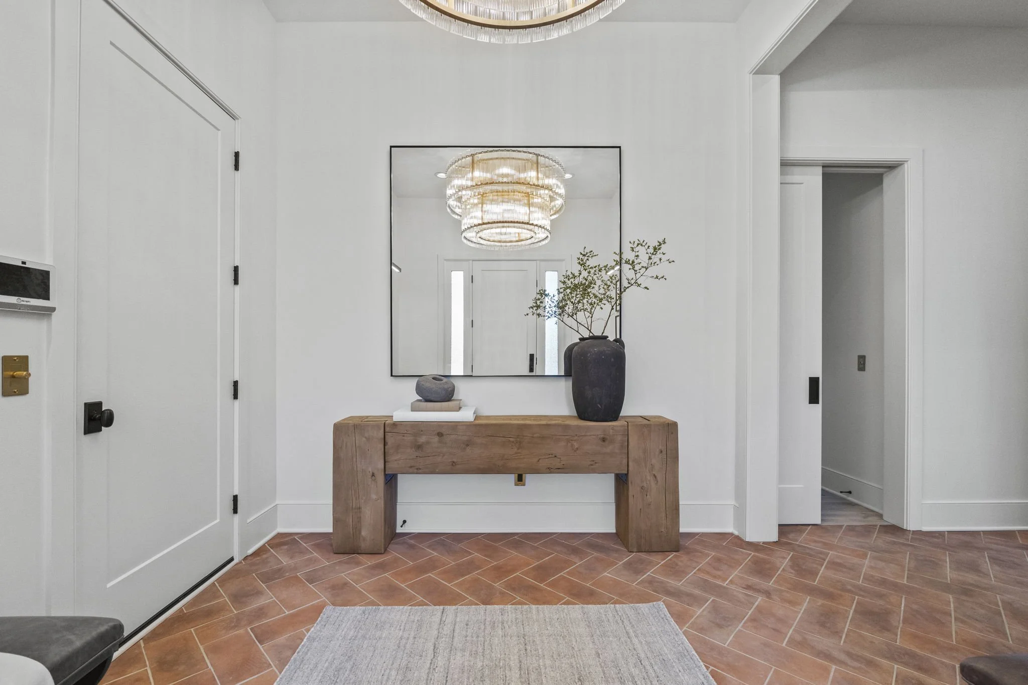 Modern entryway with a large mirror above a wooden console table, decorative vase with greenery, and a stone sculpture, illuminated by a chandelier with a layered design.