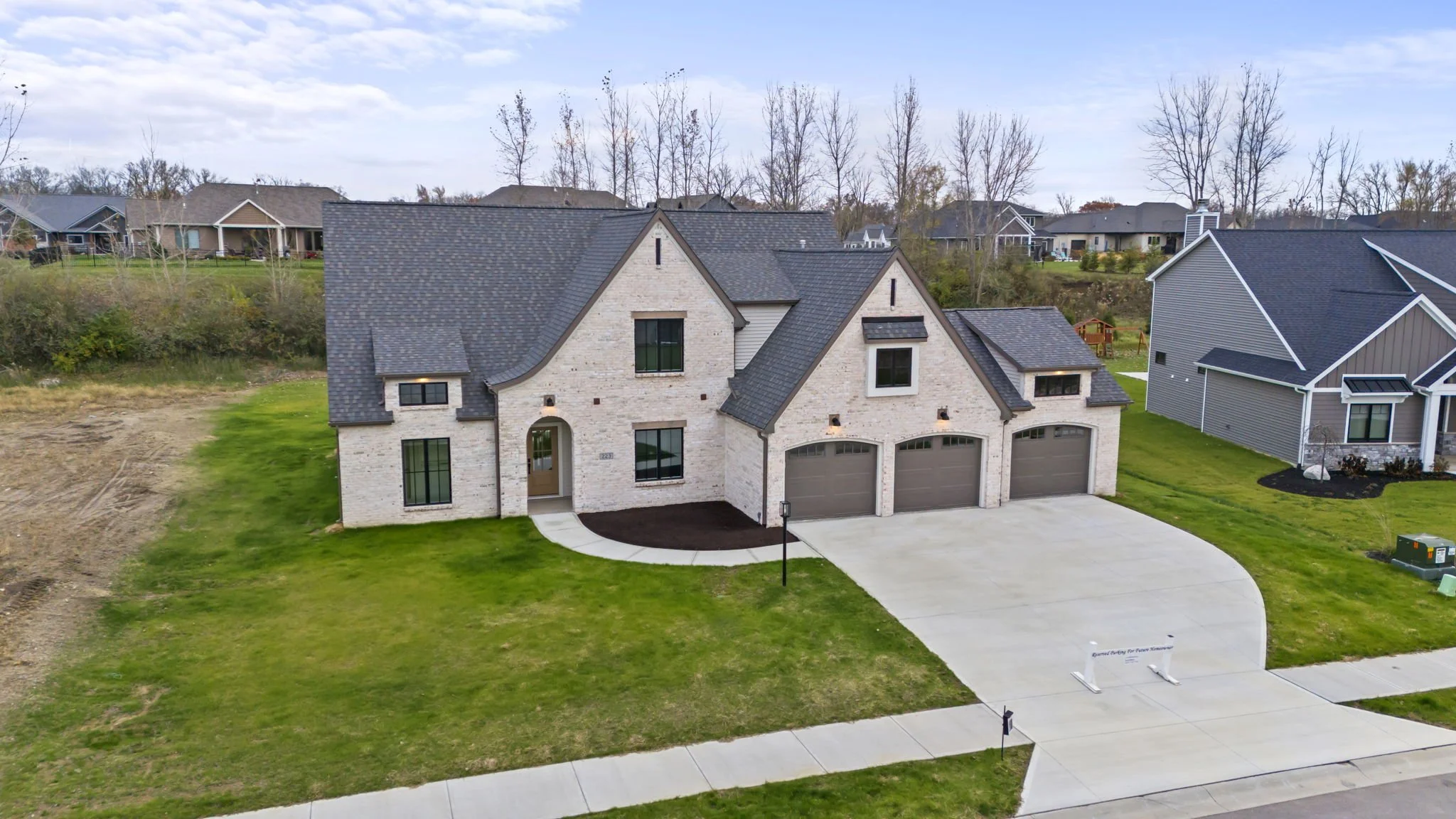 A newly constructed two-story house with a brick exterior, dark gray roof, three garage doors, a curved driveway, and a small front lawn with a sidewalk.