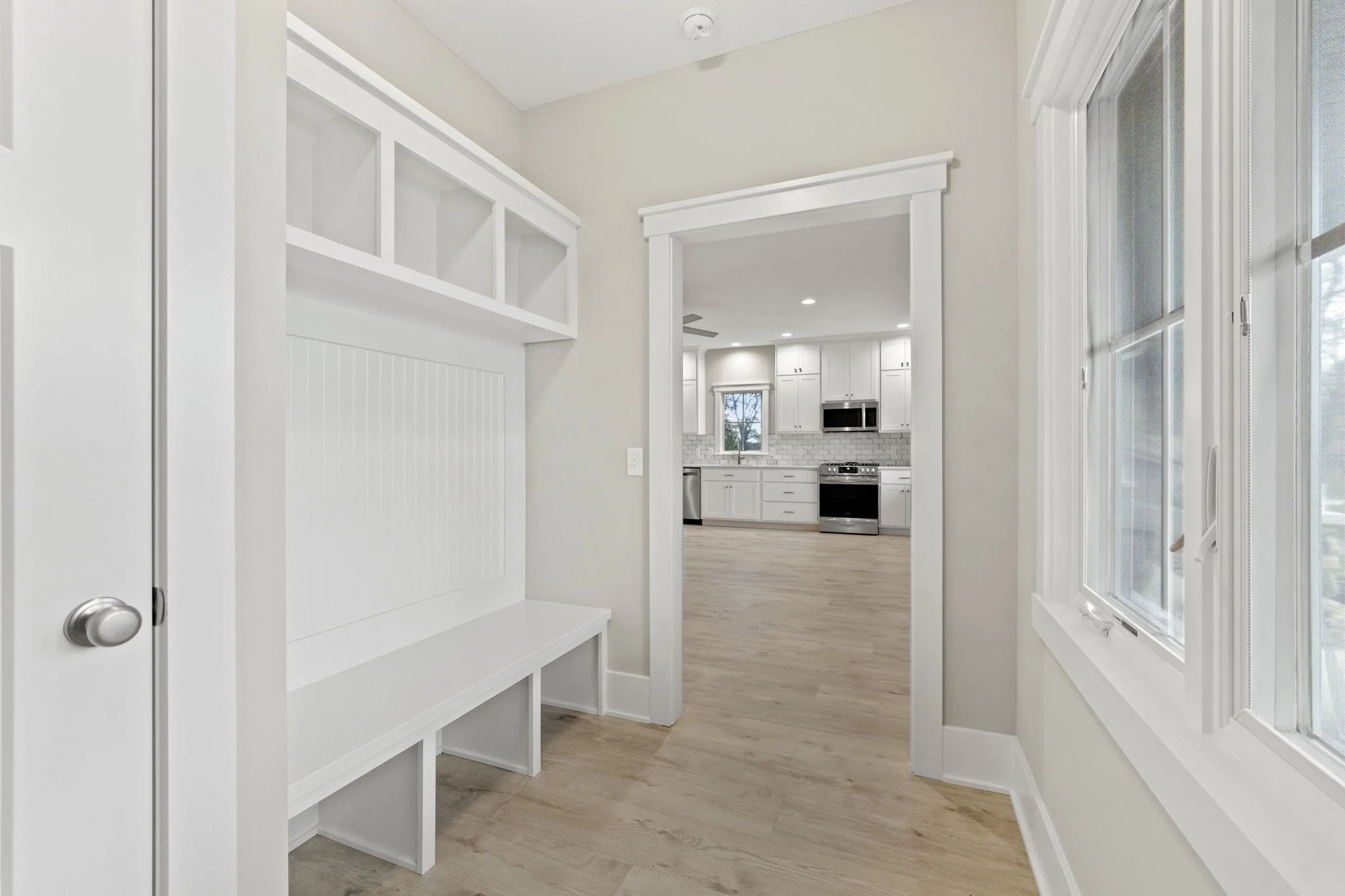View from a hallway into a kitchen with white cabinets, a window, and modern appliances, with light wood flooring and white walls.