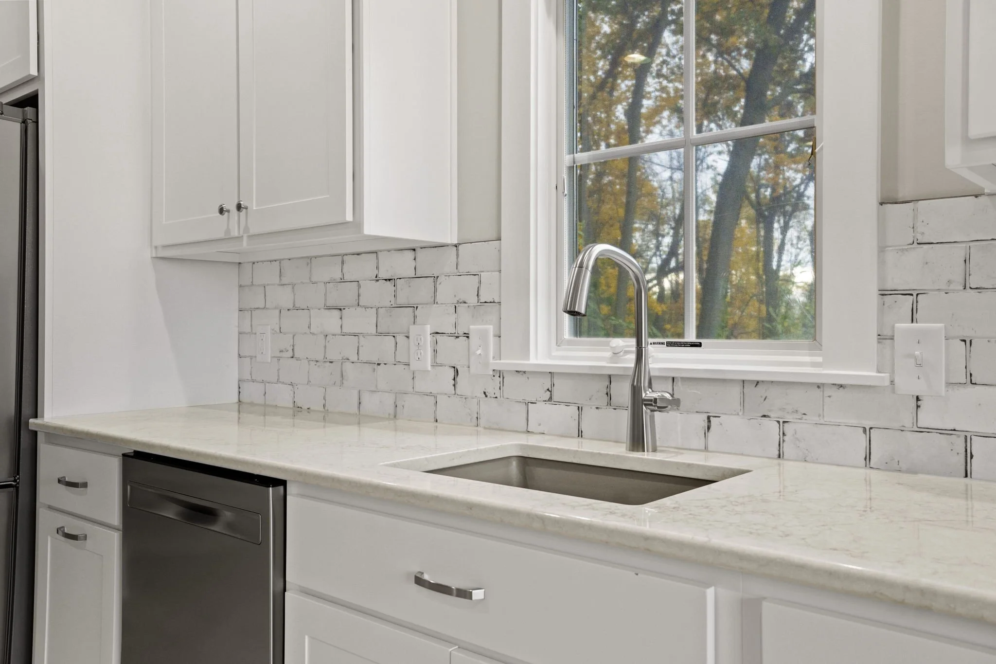 A kitchen countertop with a stainless steel sink and a chrome faucet. White cabinets above and a white brick backsplash. A window with a view of trees outside.