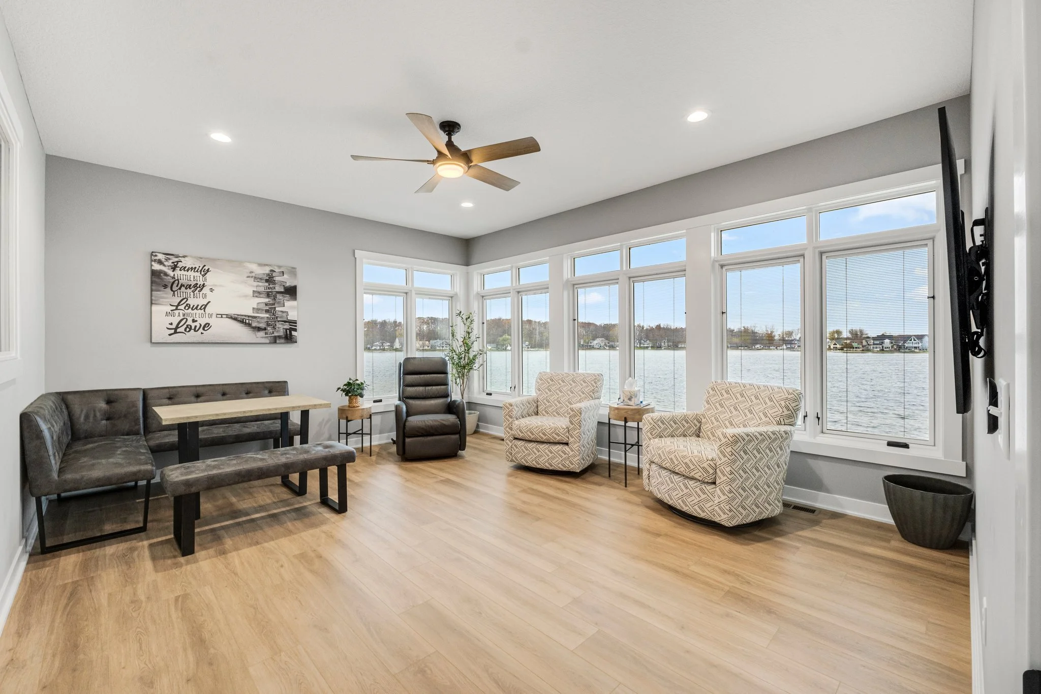 Living room with large windows overlooking water, featuring a ceiling fan, gray wall, dark gray and patterned armchairs, a black recliner, small side tables, and wall-mounted TV.