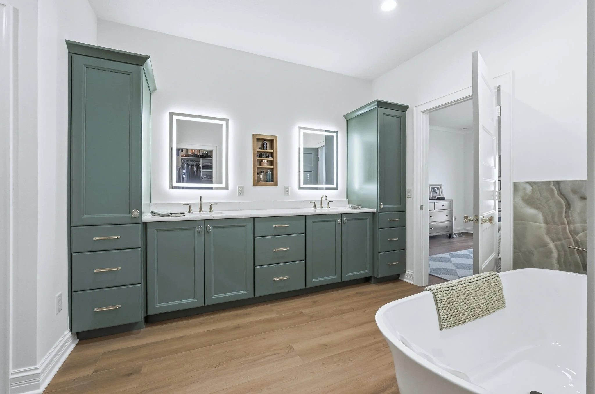 Modern bathroom with teal-colored cabinets, white countertops, two illuminated mirrors, and a white bathtub with a towel draped over the edge.