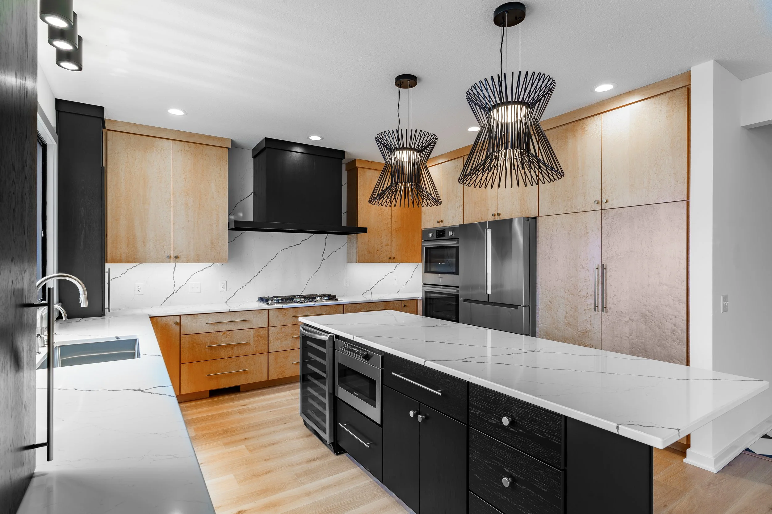 Modern kitchen with black and natural wood cabinets, a marble island, stainless steel appliances, and black wire pendant lights.