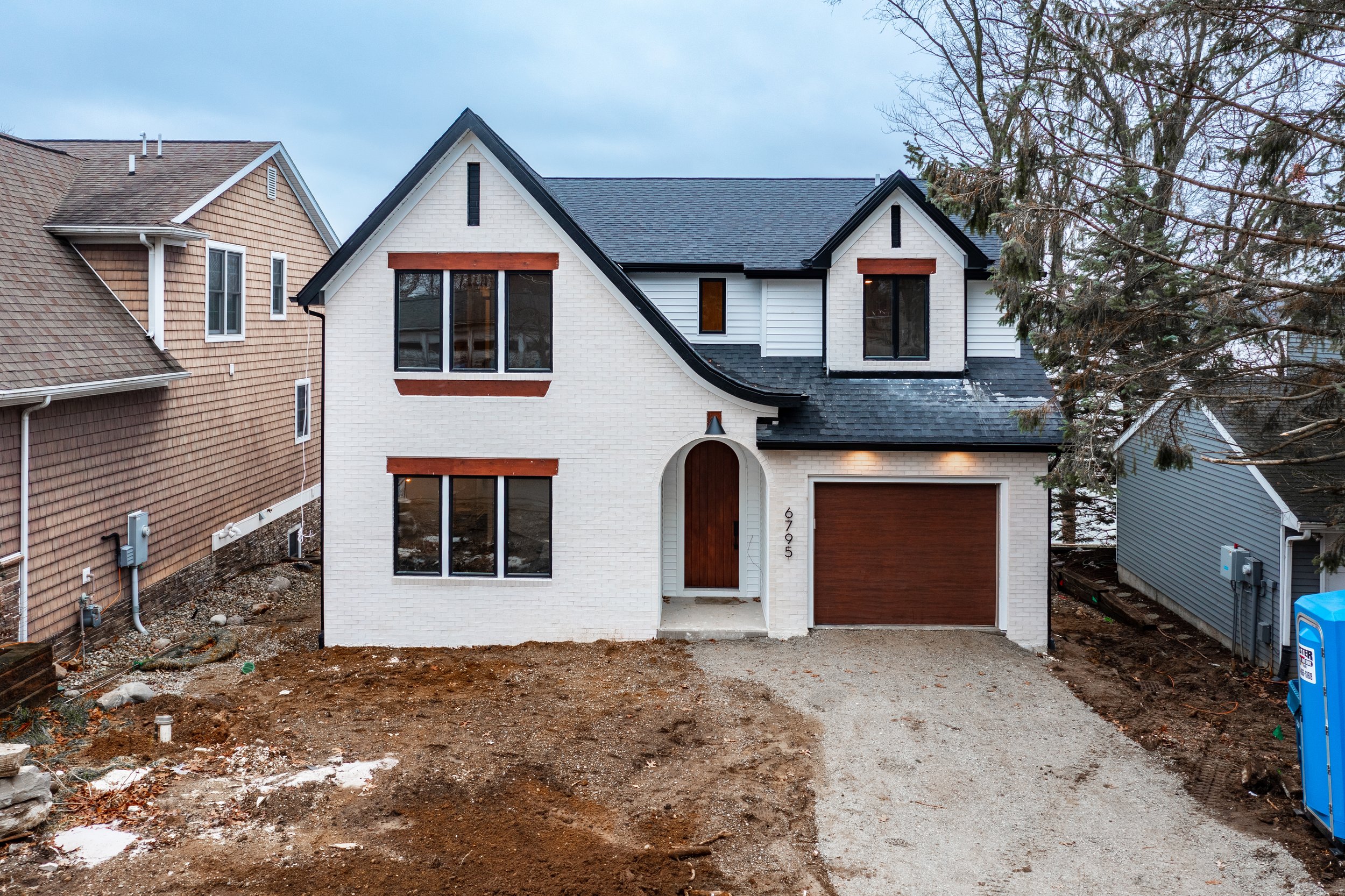 New white two-story house with a dark gray roof, brown garage door, and front door, situated between two other homes. The house has large front windows with brown trim and some construction debris and dirt in the front yard.