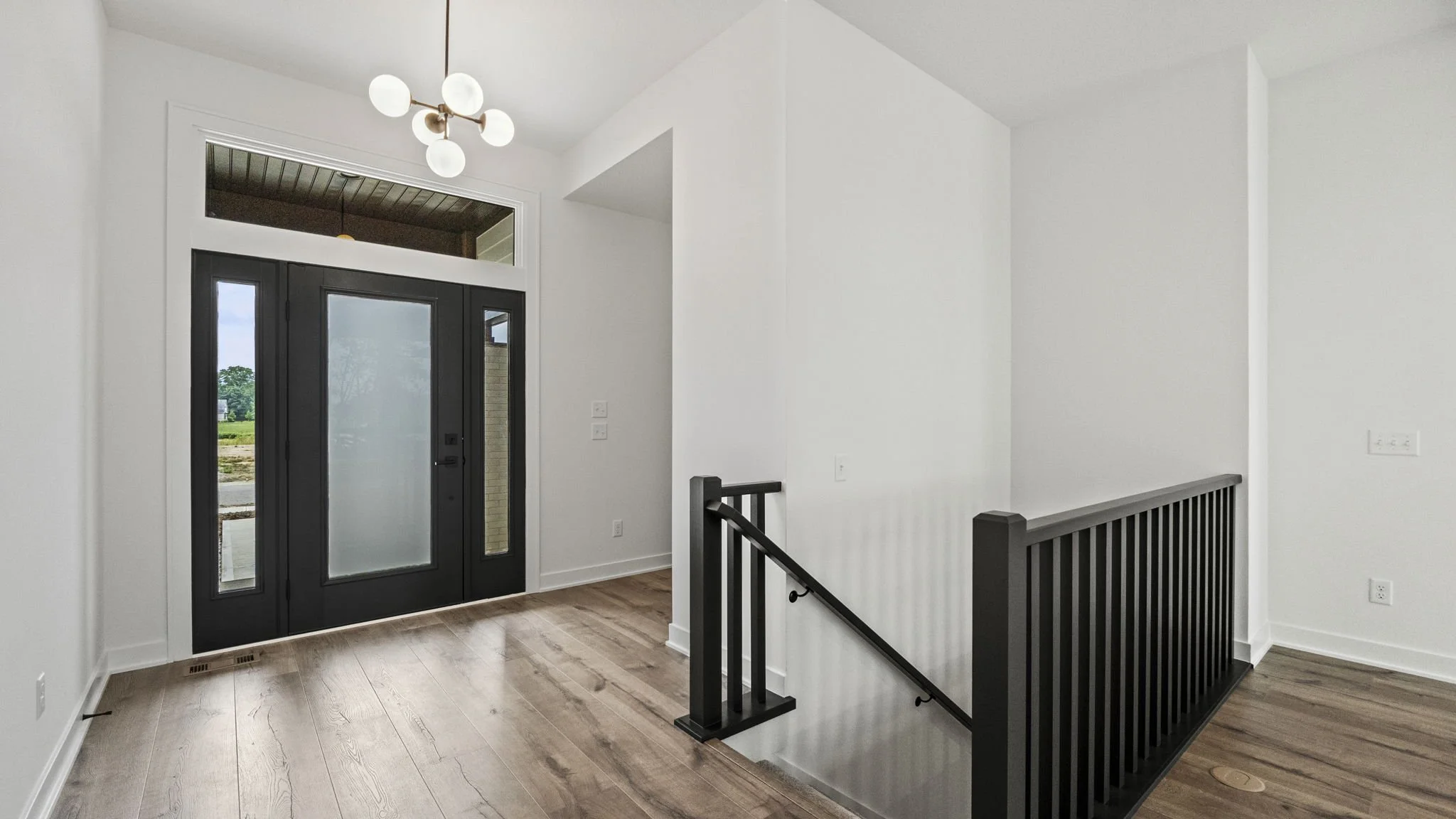Interior view of a modern house entrance with black front door, wooden flooring, white walls, and a black railing by the staircase.