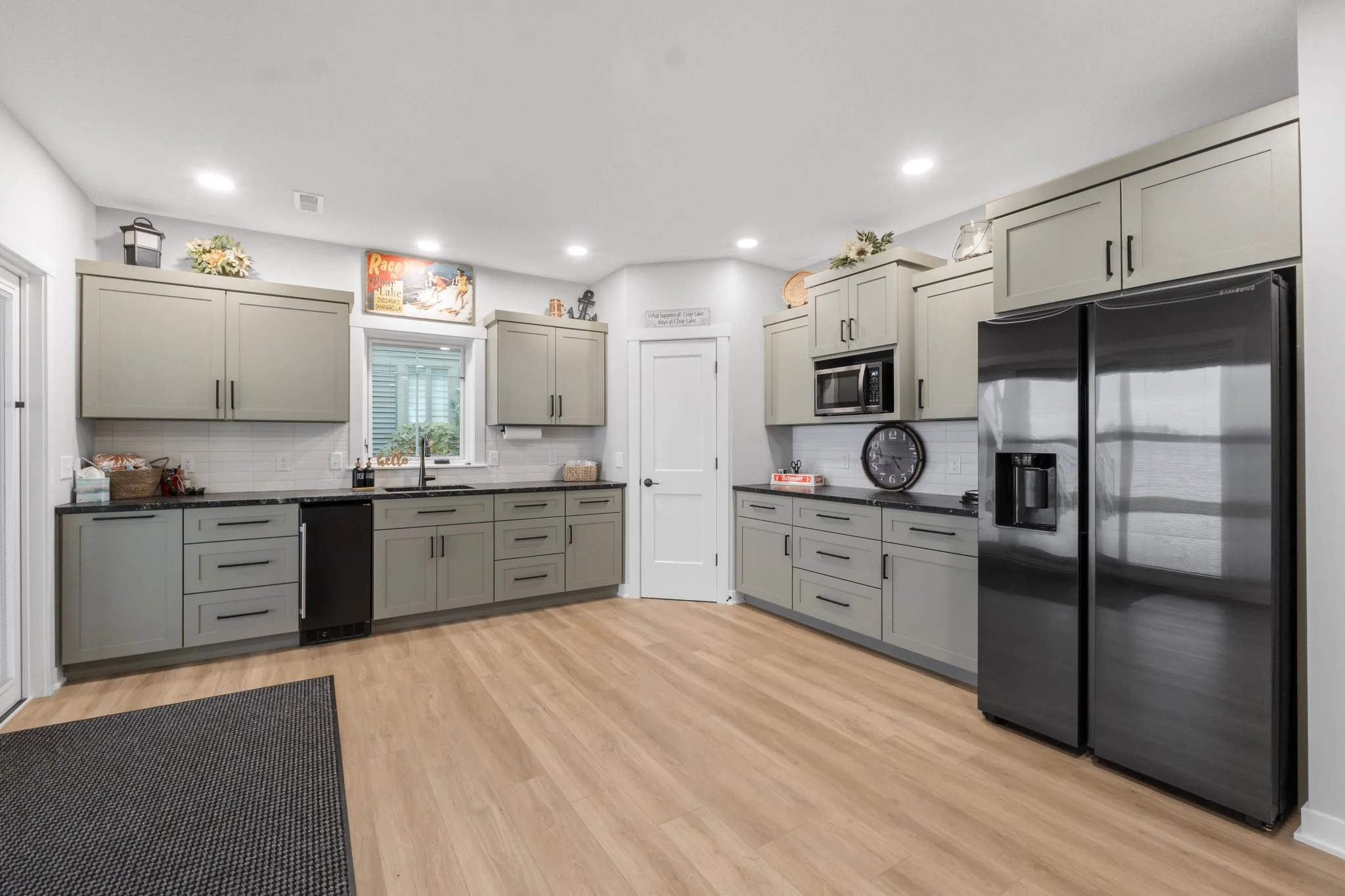 Modern kitchen with gray cabinets, black countertops, stainless steel appliances, a window above the sink, decorated with plants and signs, and wood flooring.