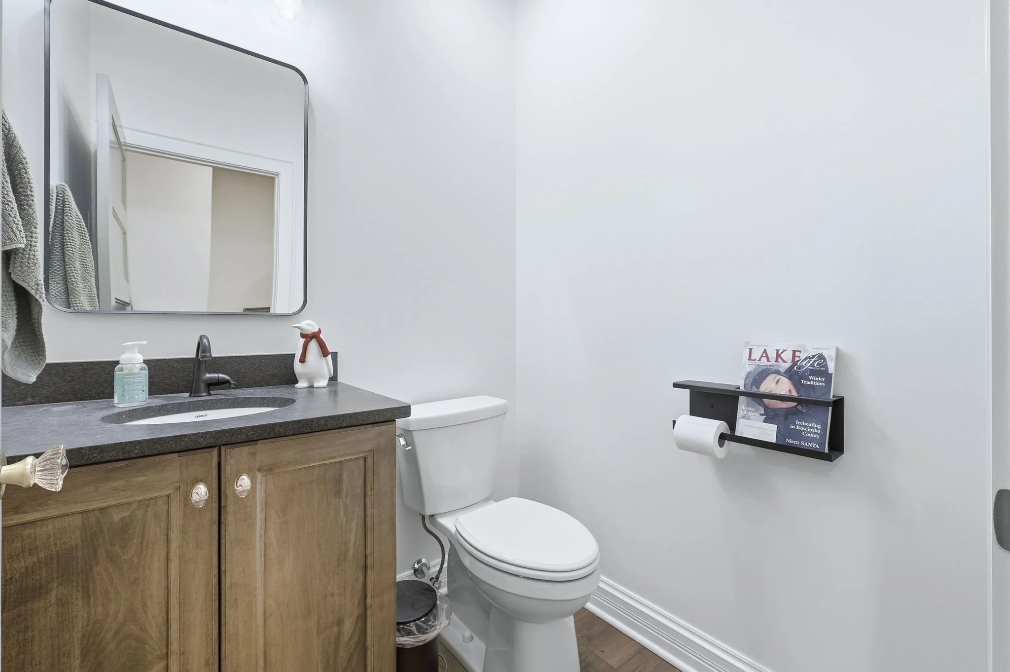 Bathroom with a wooden vanity, gray countertop, black faucet, soap dispenser, mirror, white toilet, wall magazine rack, and a roll of toilet paper.