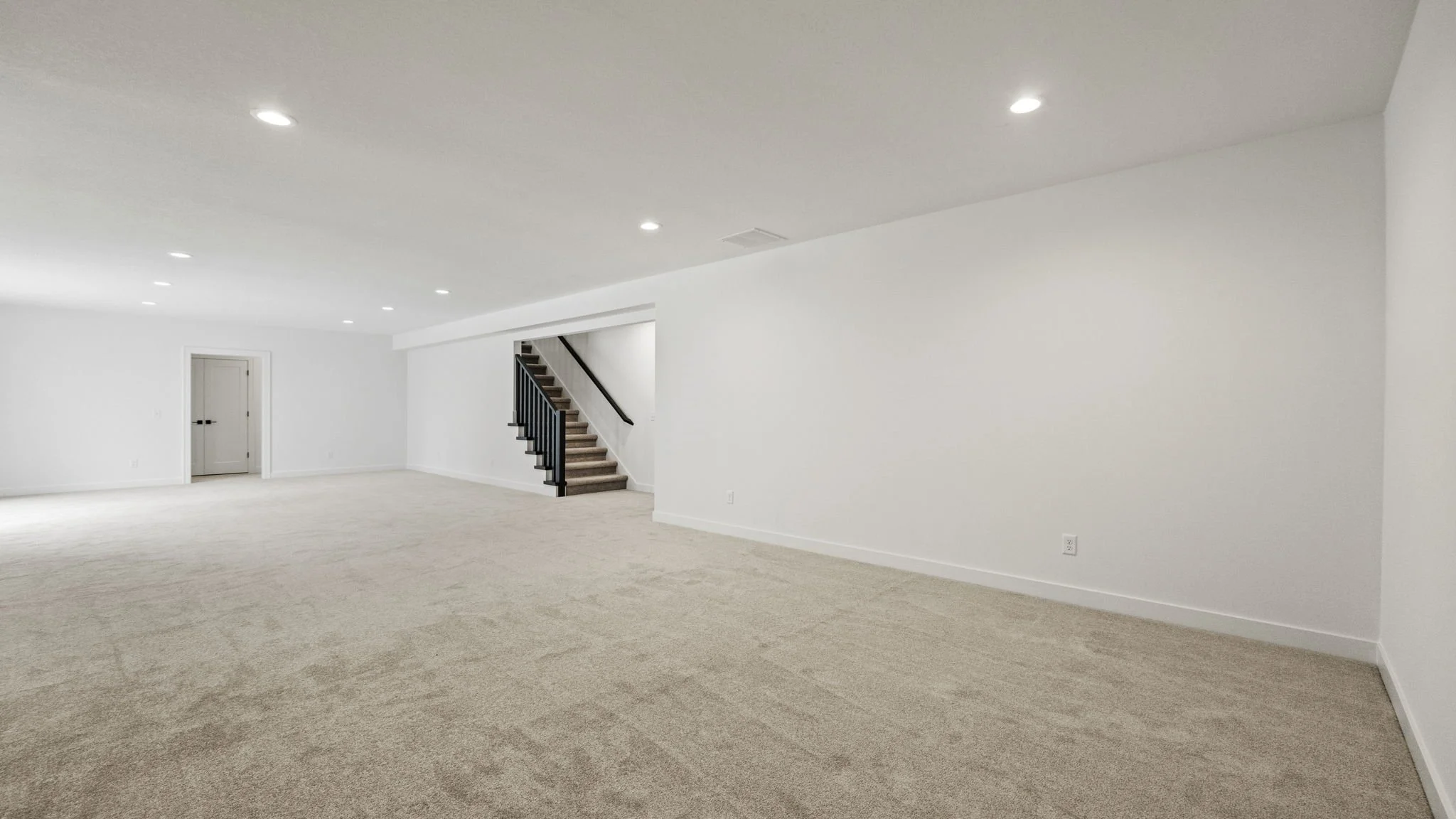Empty room with white walls, beige carpet, and ceiling with recessed lighting, staircase with black railing, and a small door on the far wall.