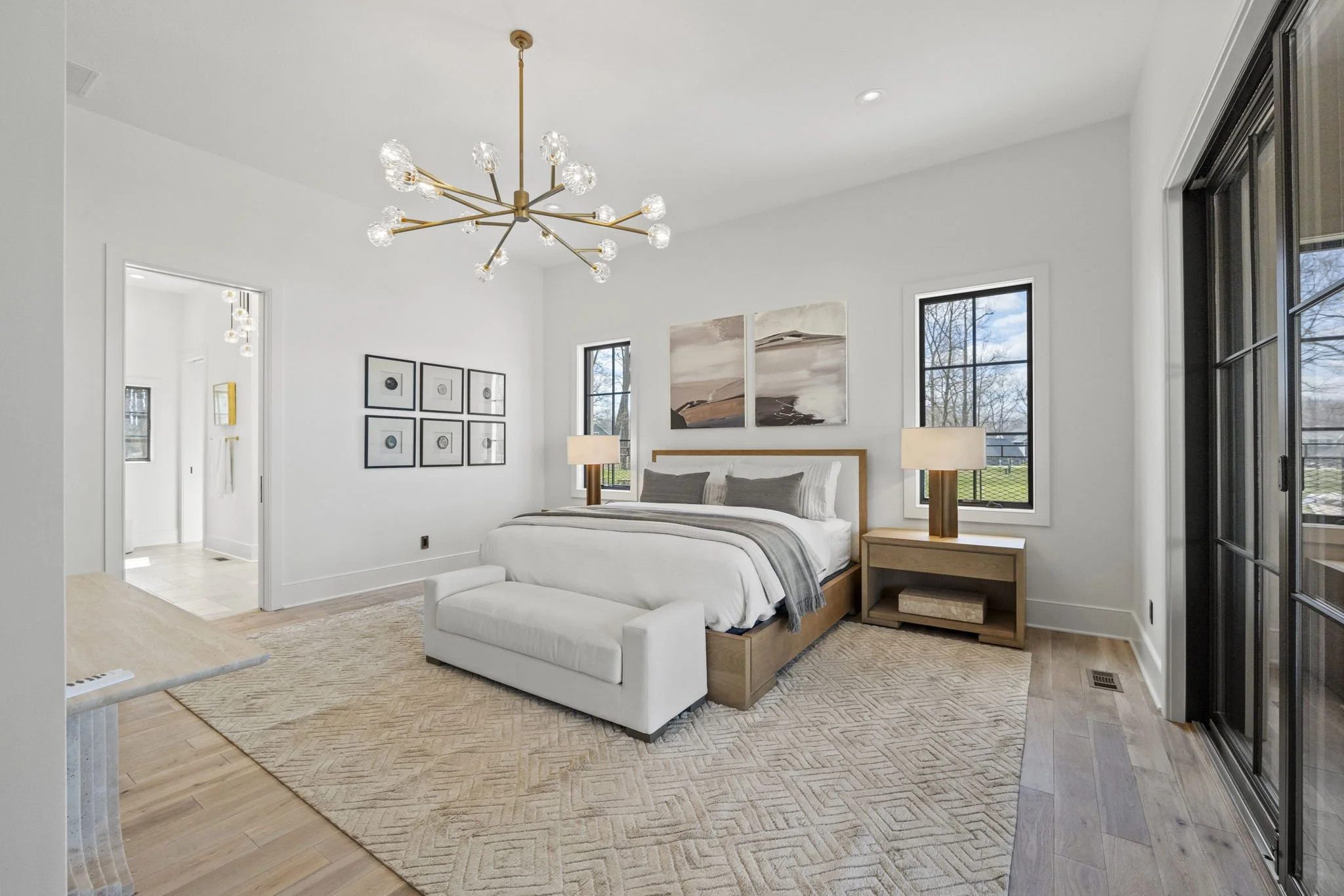 Modern bedroom with white walls, large bed, two wooden nightstands with lamps, abstract art above the bed, two vertical windows, a beige area rug, and a chandelier.