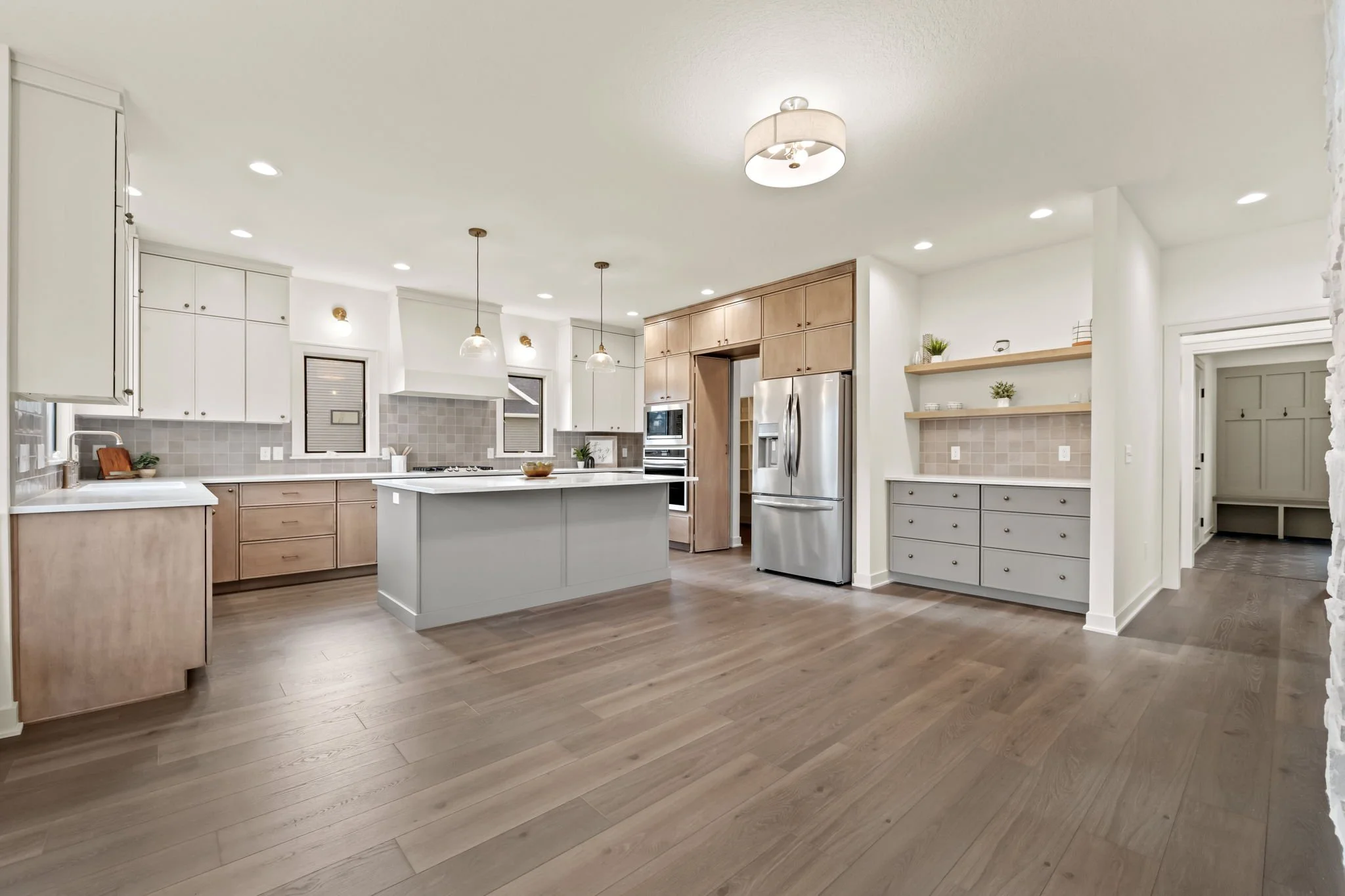 Modern kitchen with white and natural wood cabinets, stainless steel refrigerator, light-colored wood flooring, and pendant lights over a central island.