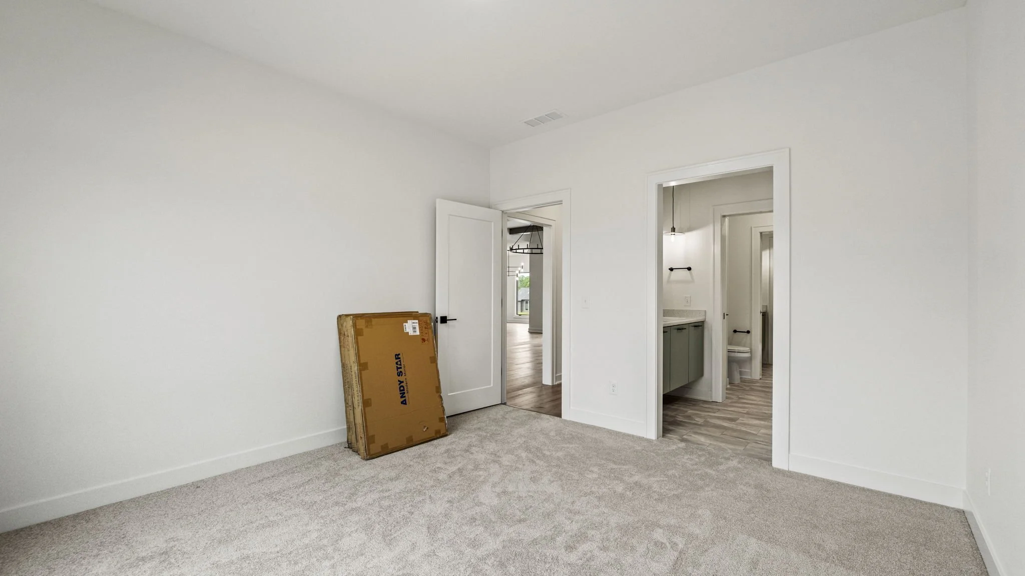Empty room with beige carpet, white walls, a partially opened door leading to another room with wooden floors, and a cardboard box leaning against the wall.