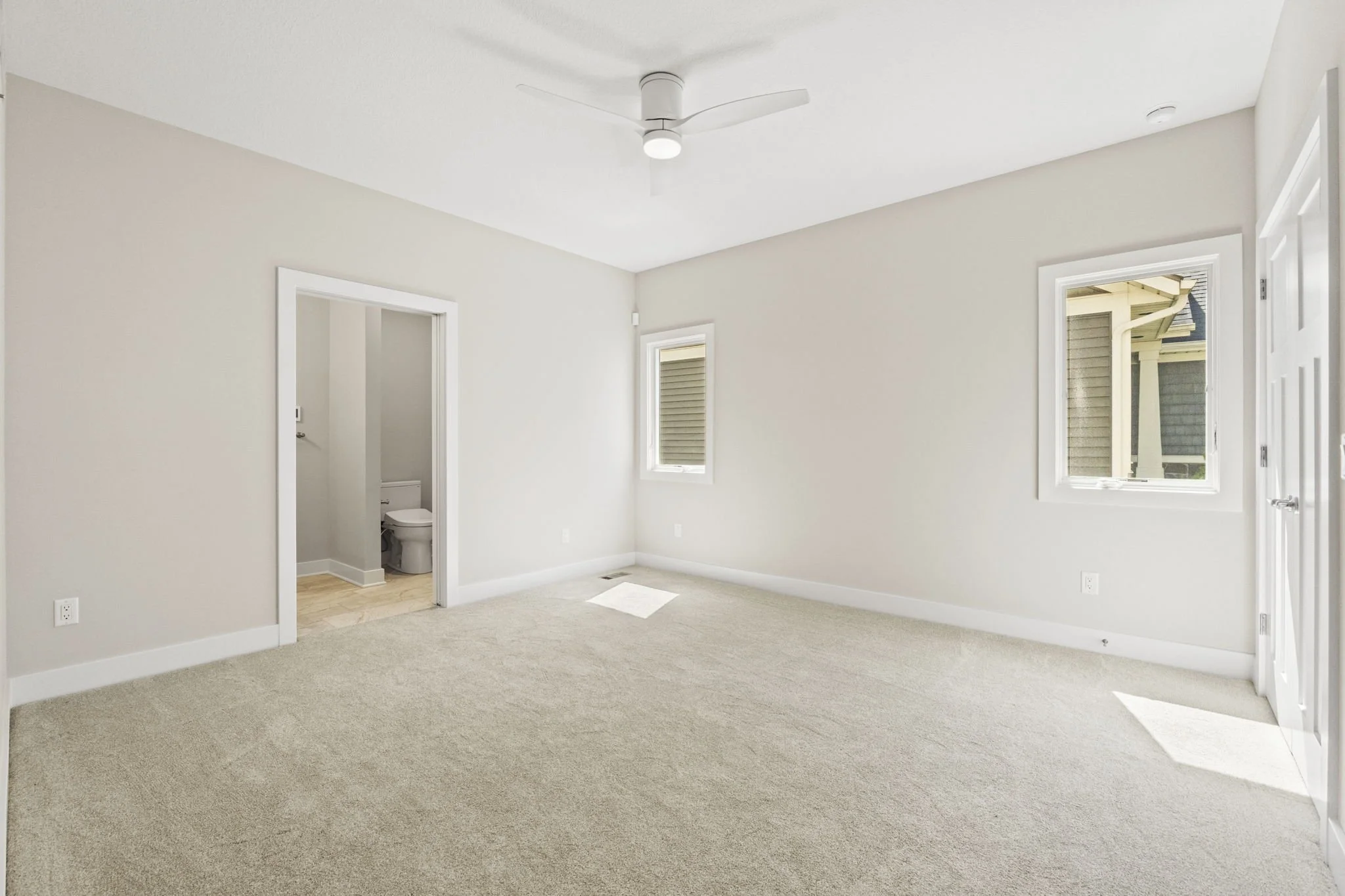 Empty bedroom with beige carpet, light-colored walls, two windows, ceiling fan, and a door to a bathroom.