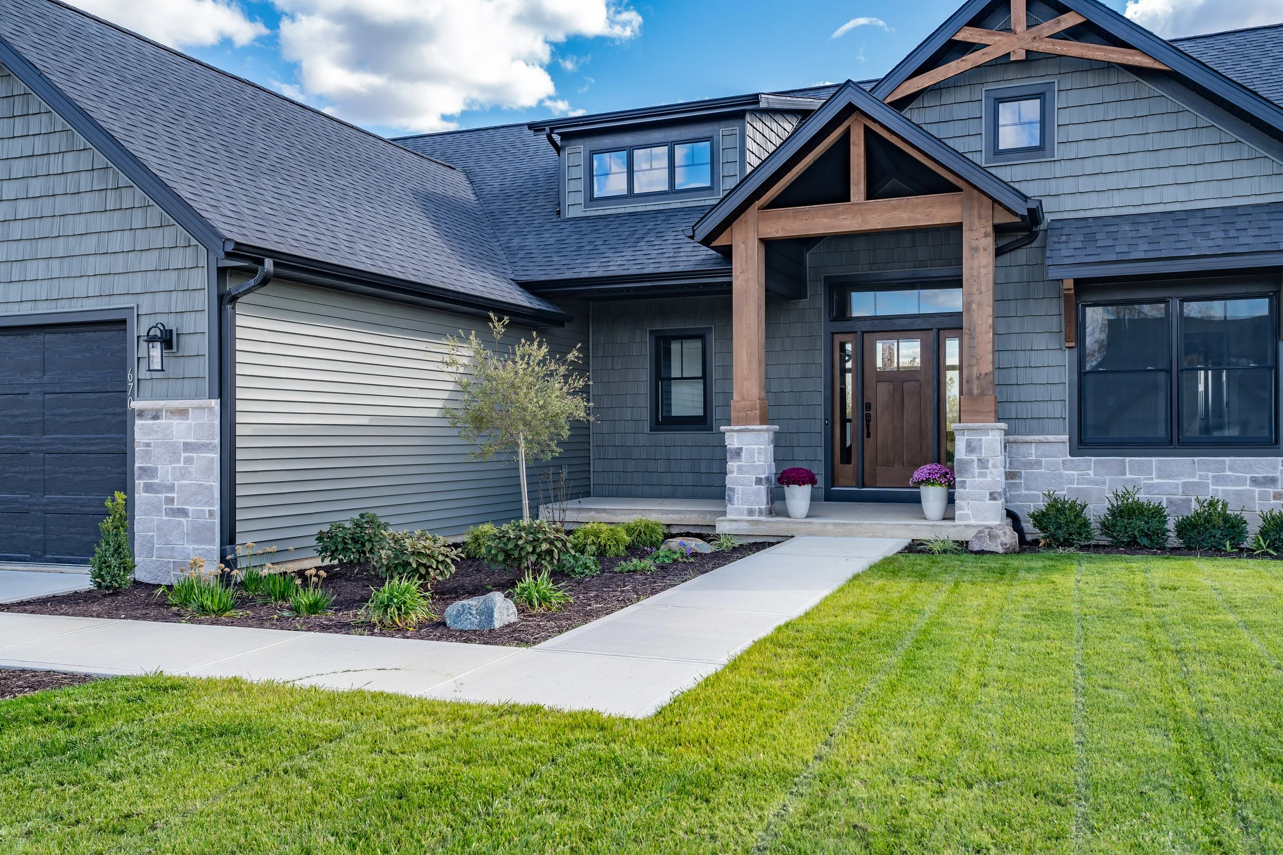 Front view of a modern house with a combination of dark gray and beige exterior, a small front porch with wooden support beams, a wooden front door, well-maintained lawn, garden beds with plants, and a concrete walkway leading to the entrance.