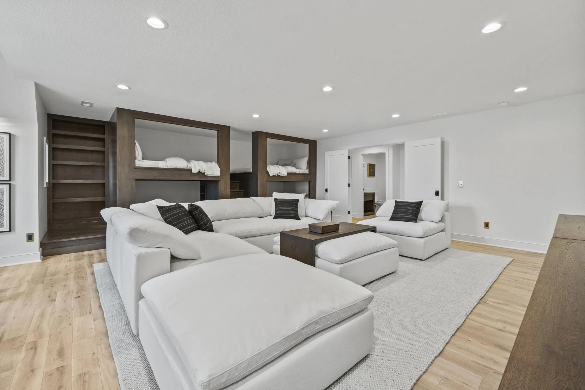 Spacious living room with white sectional sofa, black accent pillows, light wood flooring, and built-in bunk beds with dark wood frames.