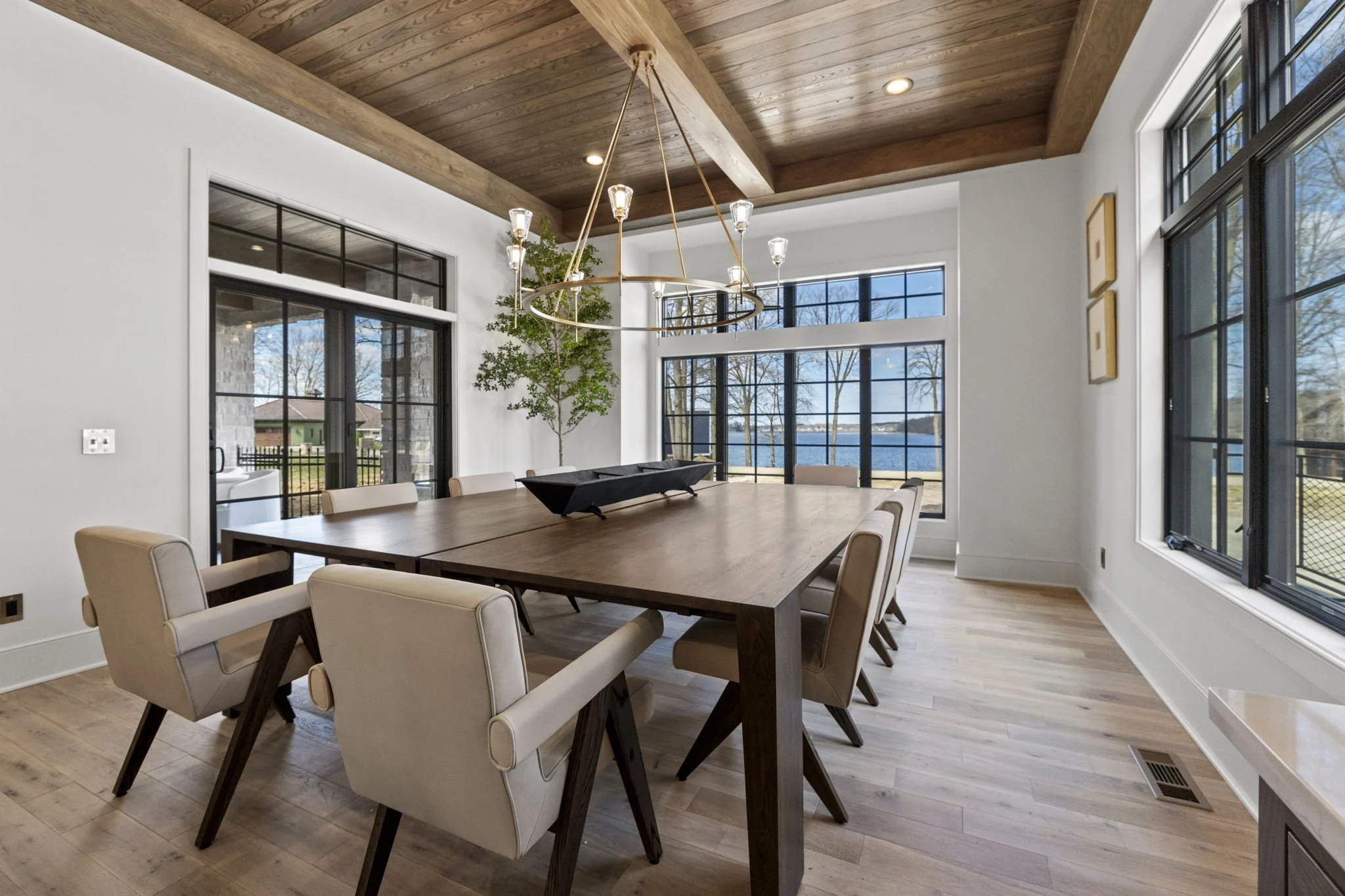 Modern dining room with a wooden table, beige chairs, large windows, and a view of water outside.