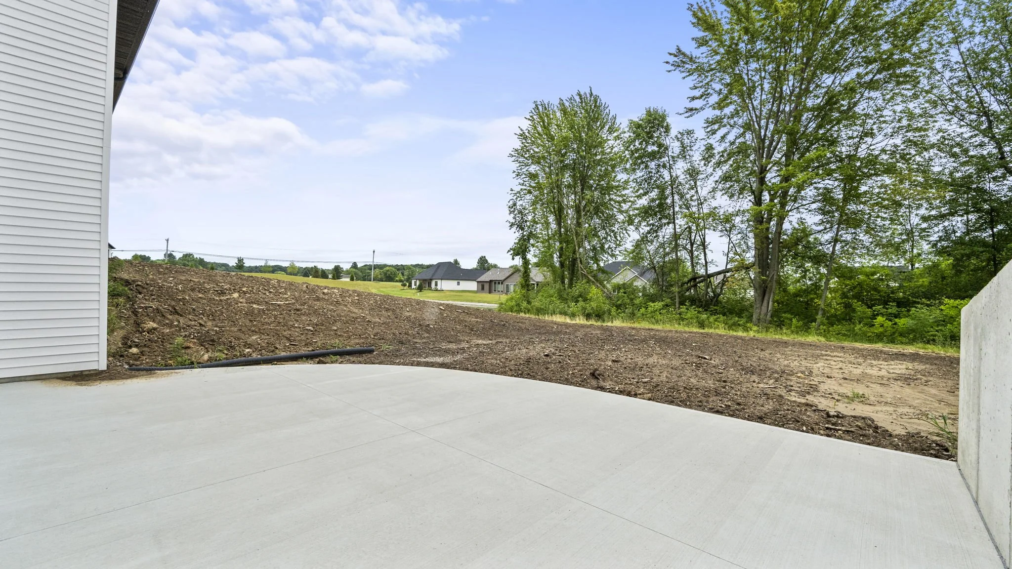 View from a concrete patio showing a dirt yard with some trees and neighboring houses in the distance under a partly cloudy sky.