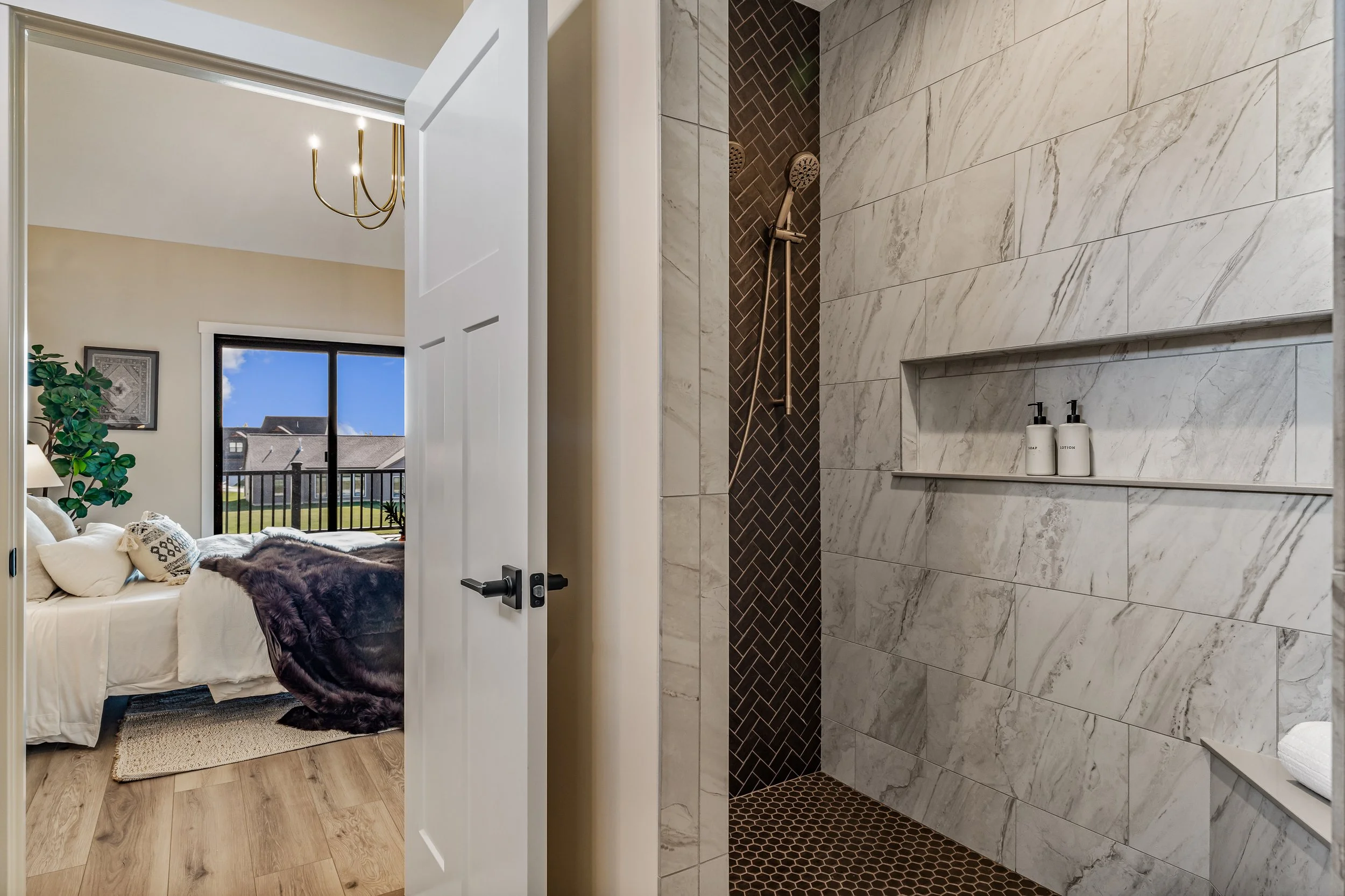 View from bathroom into bedroom showing a walk-in shower with white marble tiles and dark accent tiles, a bedroom with a bed, pillows, dark blanket, window with a view of houses and blue sky.