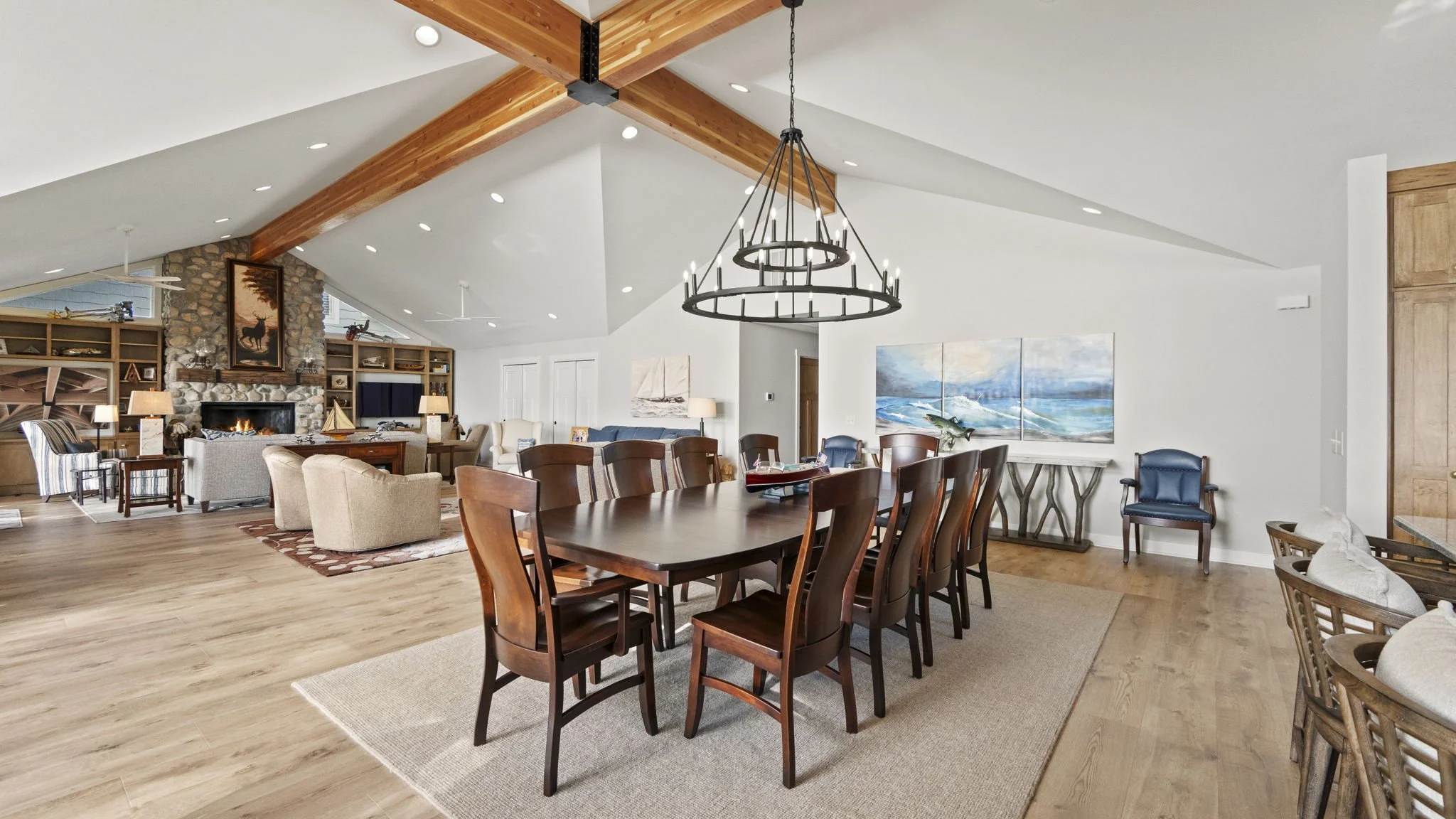 Open-concept living and dining room in a modern home. Features a large wooden dining table with eight chairs, a cozy seating area with armchairs and a stone fireplace, wooden beams on the ceiling, and artwork on the walls. Light wood flooring and neu