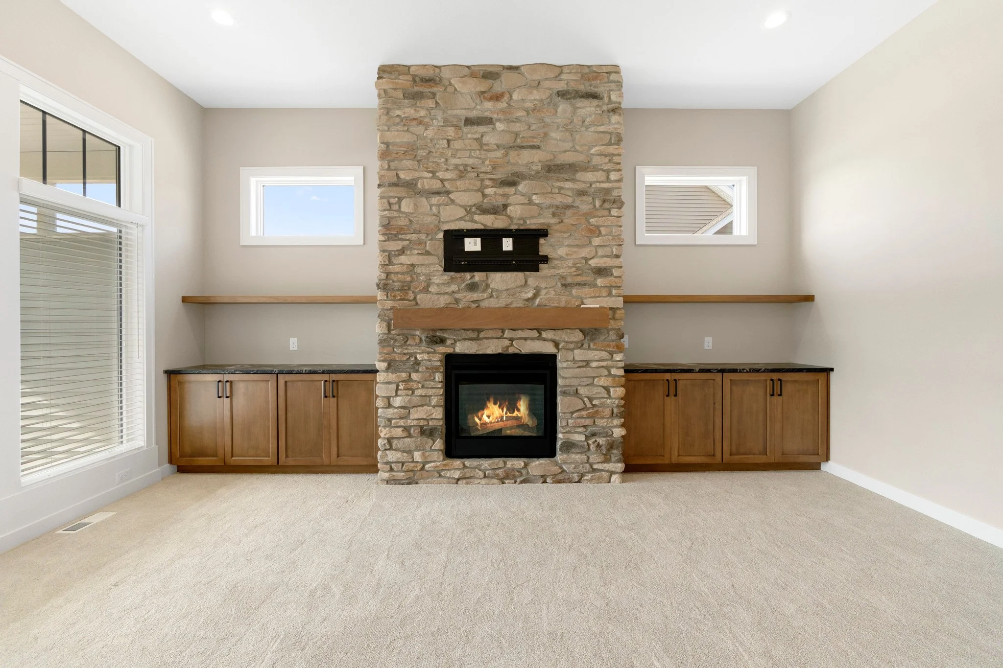 Living room with a central stone fireplace, two small windows above, wooden cabinetry along the lower walls, and beige carpet flooring.