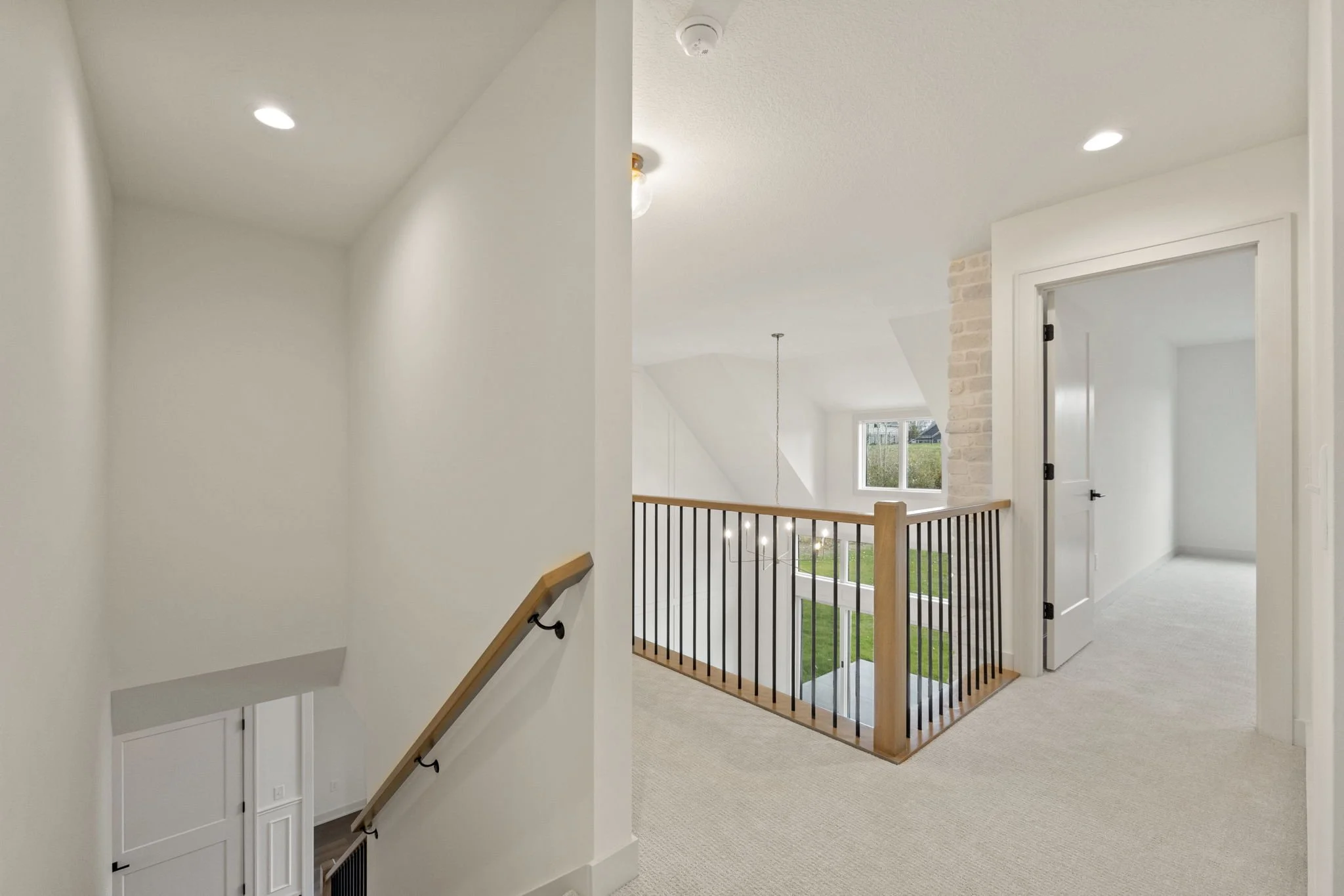Bright, white upstairs hallway with carpeted flooring, a staircase with a wooden handrail and black balusters, and a door leading to a room. Large window overlooking a green yard, brick wall accents, and ceiling lights.