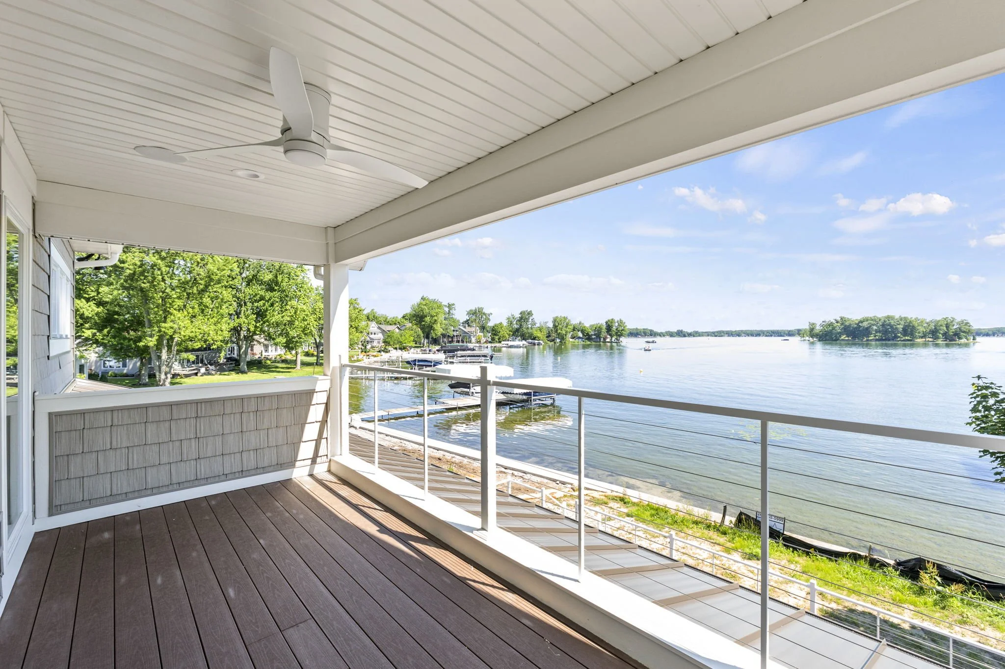 View from a covered balcony overlooking a waterfront with a beach, trees, boats, and a small island in the distance on a clear sunny day.