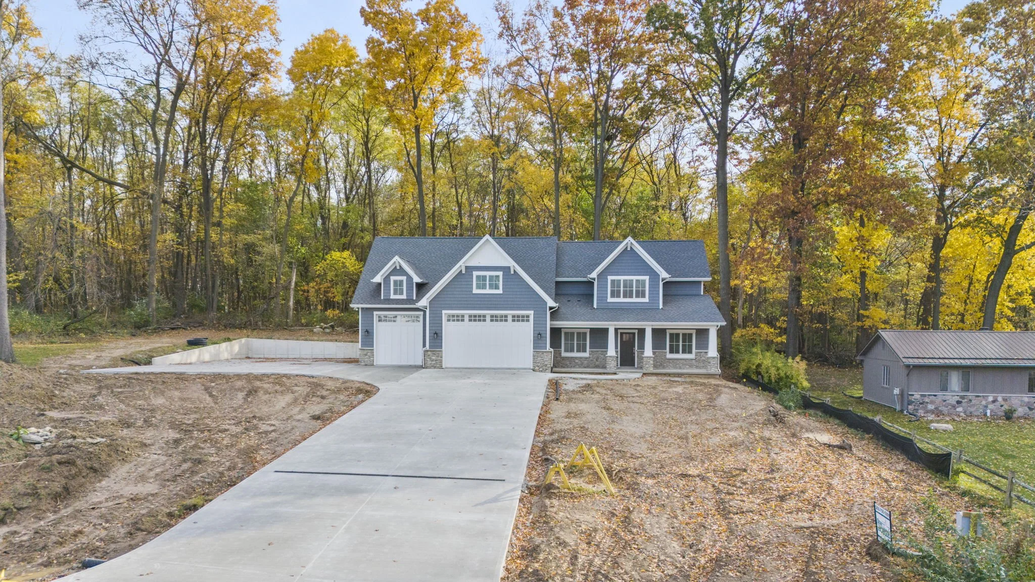 Newly constructed two-story house with gray siding and white trim, two garage doors, situated in a wooded area during fall with leaves changing color.