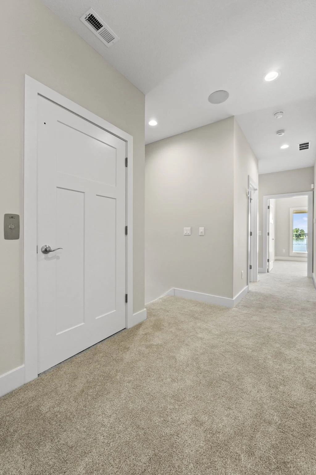 Empty hallway with white walls, beige carpet, and white doors leading to bedrooms with bright natural light.
