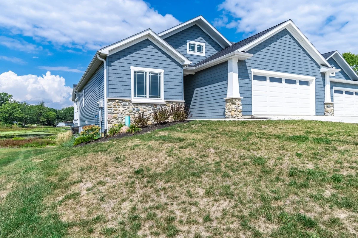Blue house with stone accents and attached garage, situated on a grassy lawn under a partly cloudy sky.