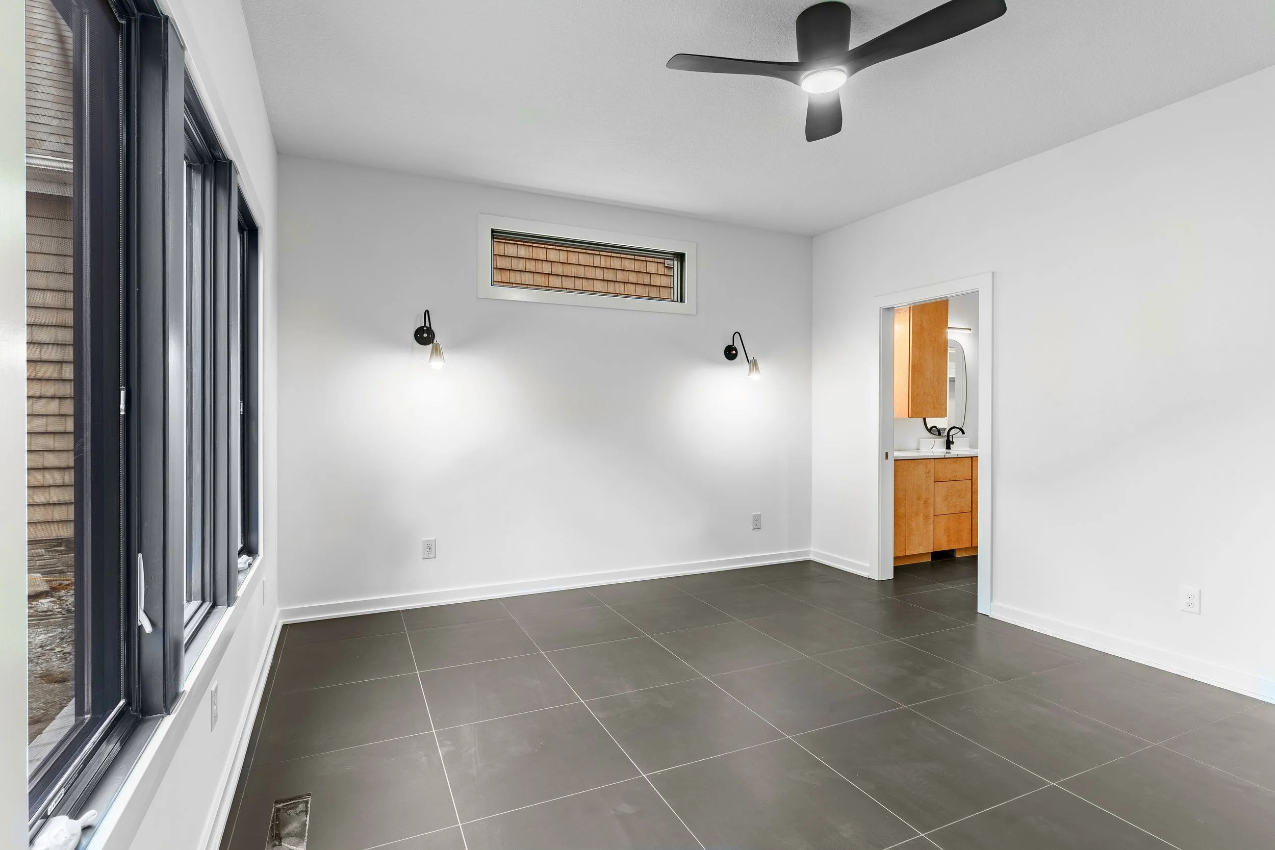 Empty room with large windows, white walls, dark tiled flooring, and a ceiling fan, with a doorway leading to a small bathroom with wooden cabinetry and a mirror.