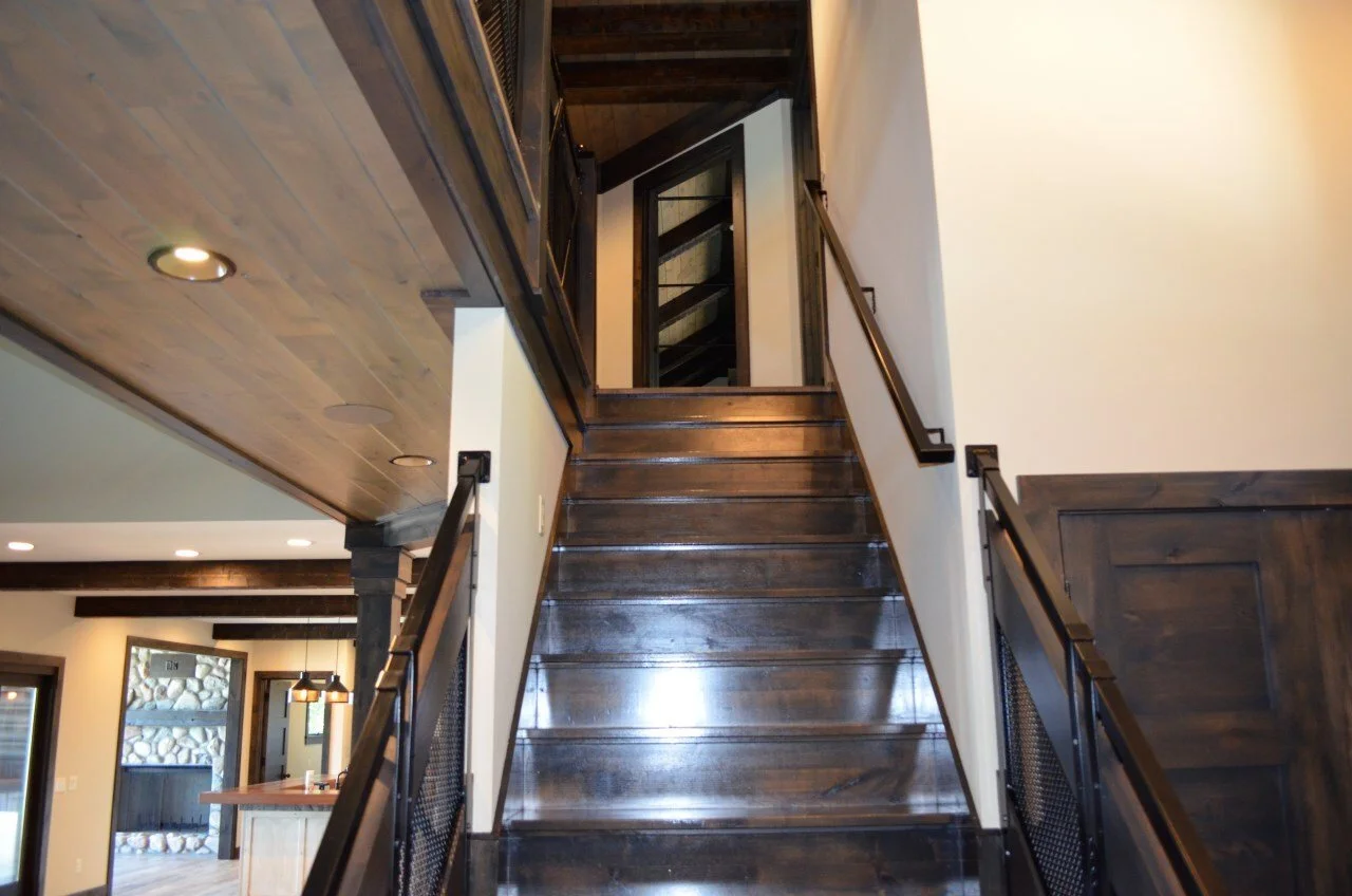Wooden staircase leading upstairs with black metal railing on both sides, in a modern home interior with wood and white walls, visible kitchen and living area in the background.