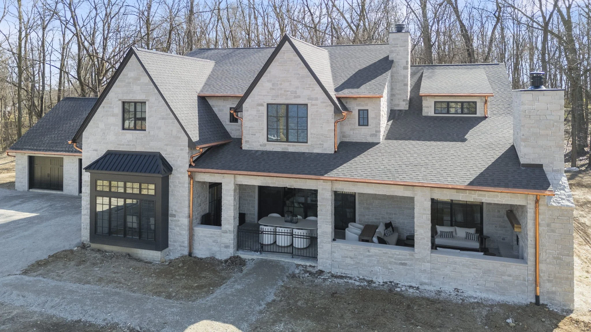 A modern two-story house with stone exterior walls, multiple gabled roofs, and large windows, surrounded by a wooded area in the background.