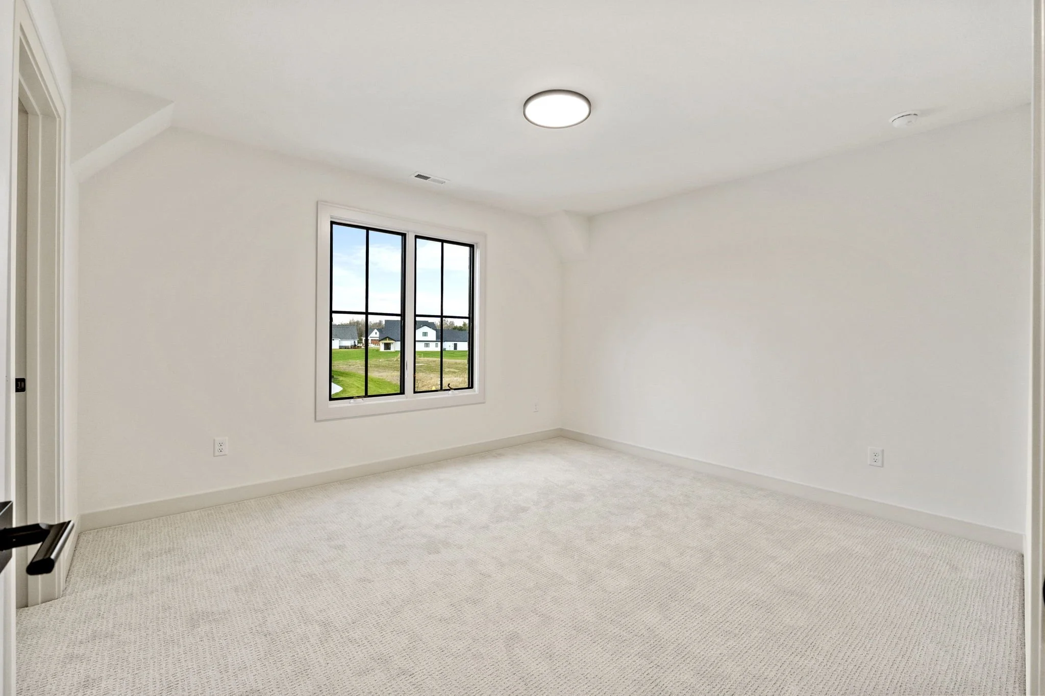 Empty white room with a window showing a view of a grassy yard and houses outside, carpeted floor, ceiling light, and electrical outlets.