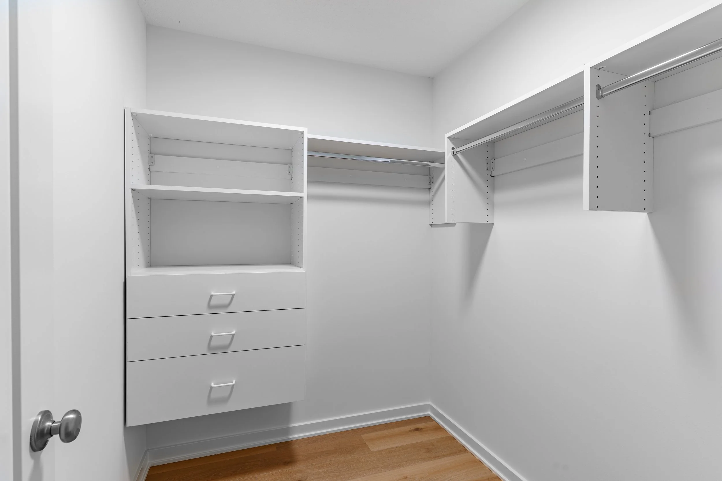 Empty white walk-in closet with built-in shelves, drawers, and hanging rods, and hardwood floor.