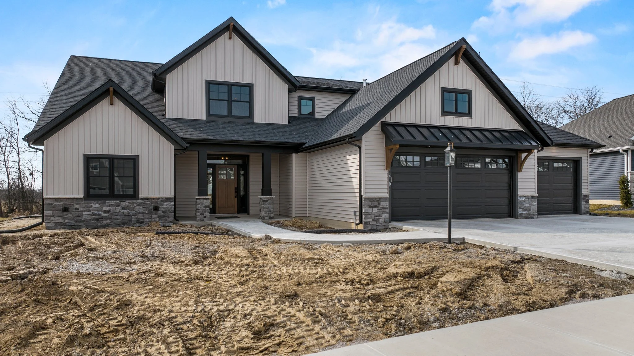 A newly constructed house with a two-car garage, dark gray doors, beige siding, stone accents at the bottom, black window frames, and a front porch with a wooden door. The yard is unfinished with dirt and a concrete walkway leading to the entrance.