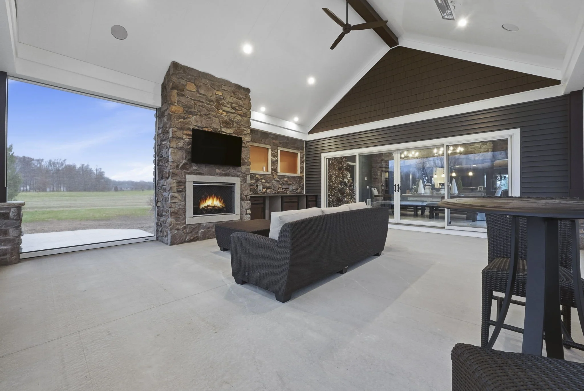 Living room with large sliding glass doors, stone fireplace, TV above fireplace, ceiling fan, white cushions on dark wicker furniture, view of a grassy field outside, tan carpet