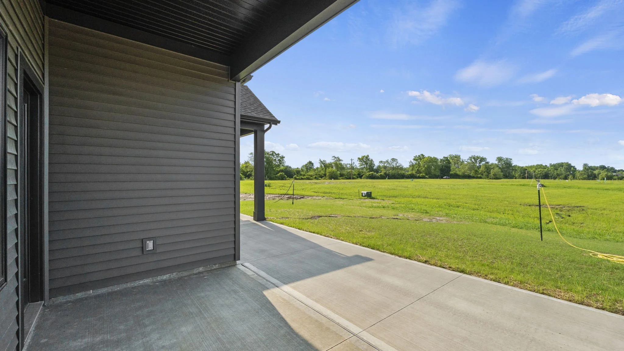 View of a backyard patio with gray siding, concrete floor, and open grassy field under a blue sky with scattered clouds.