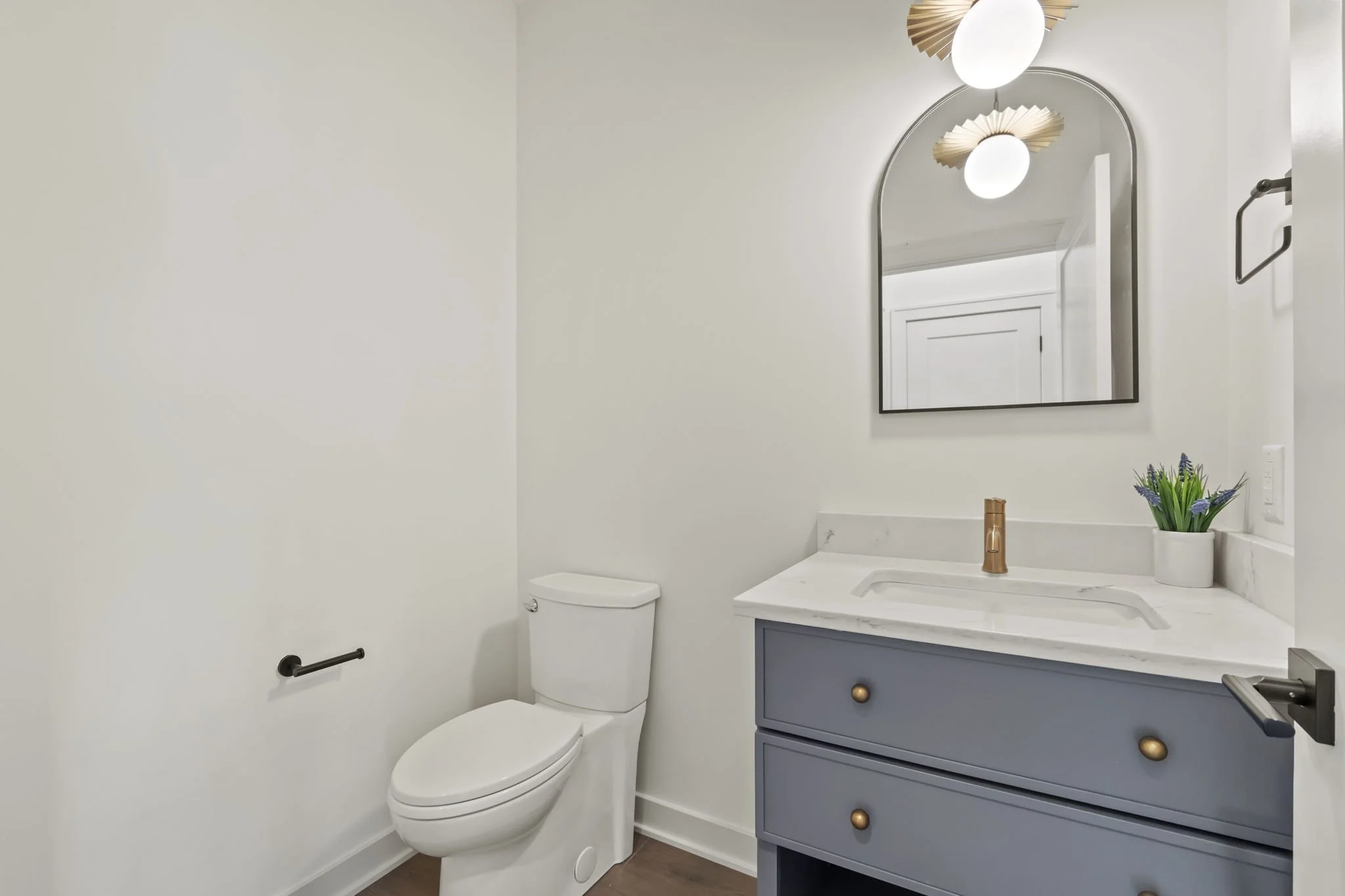 A modern bathroom with a white toilet, a blue vanity with gold knobs, a white marble countertop, a tall mirror, a potted lavender plant, and a wall-mounted towel bar.