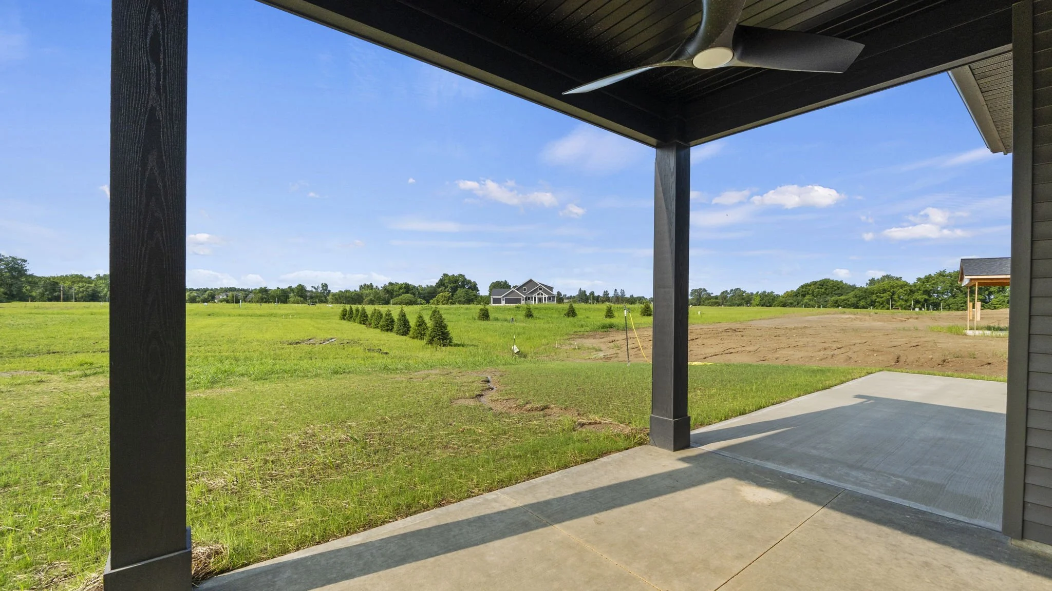 View from a covered patio showing a large open grassy field with small trees, a house in the distance, and a house under construction; blue sky with scattered clouds.