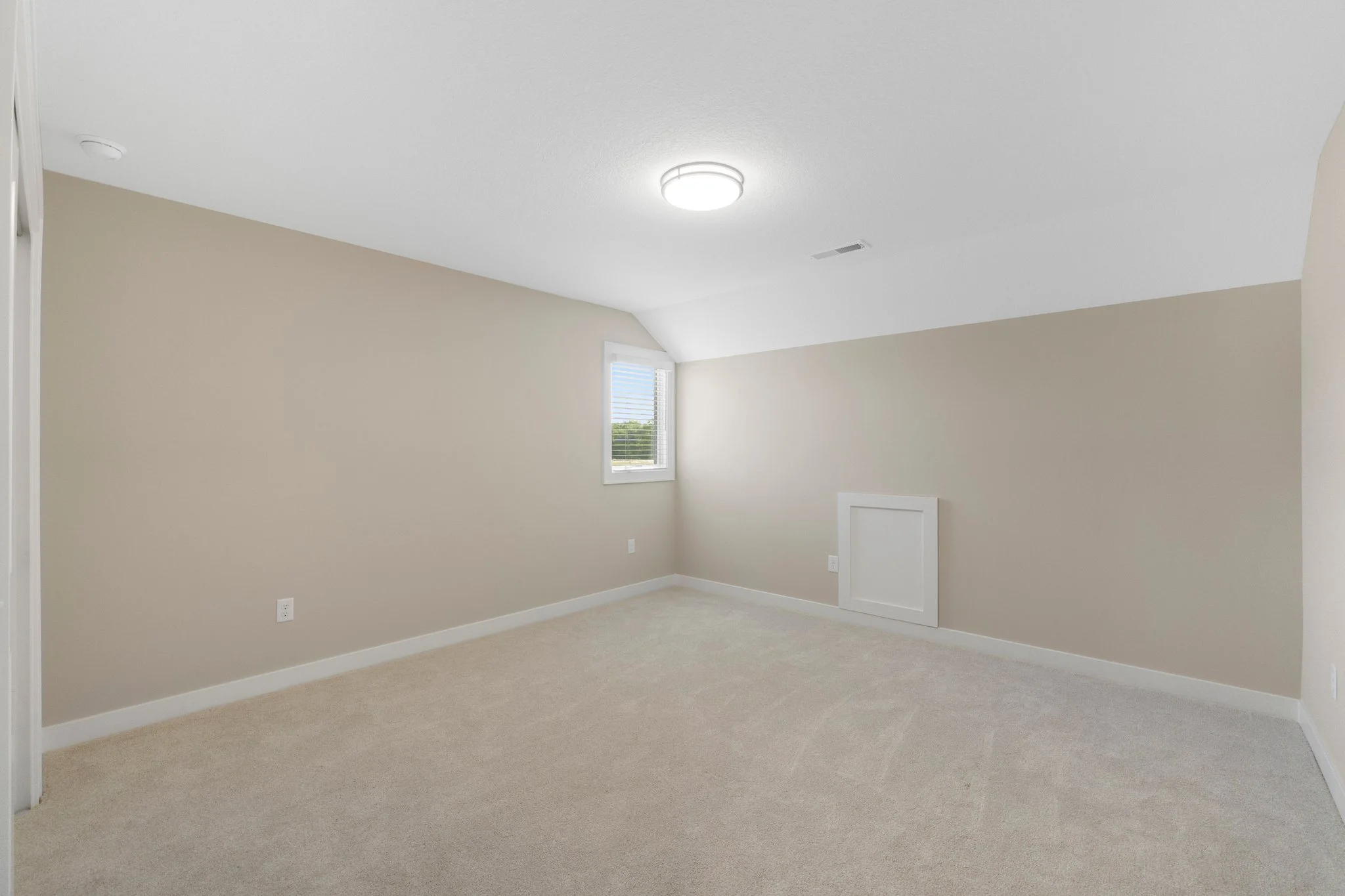 Empty beige room with a small window, a ceiling light, and a door with an access panel on the wall.