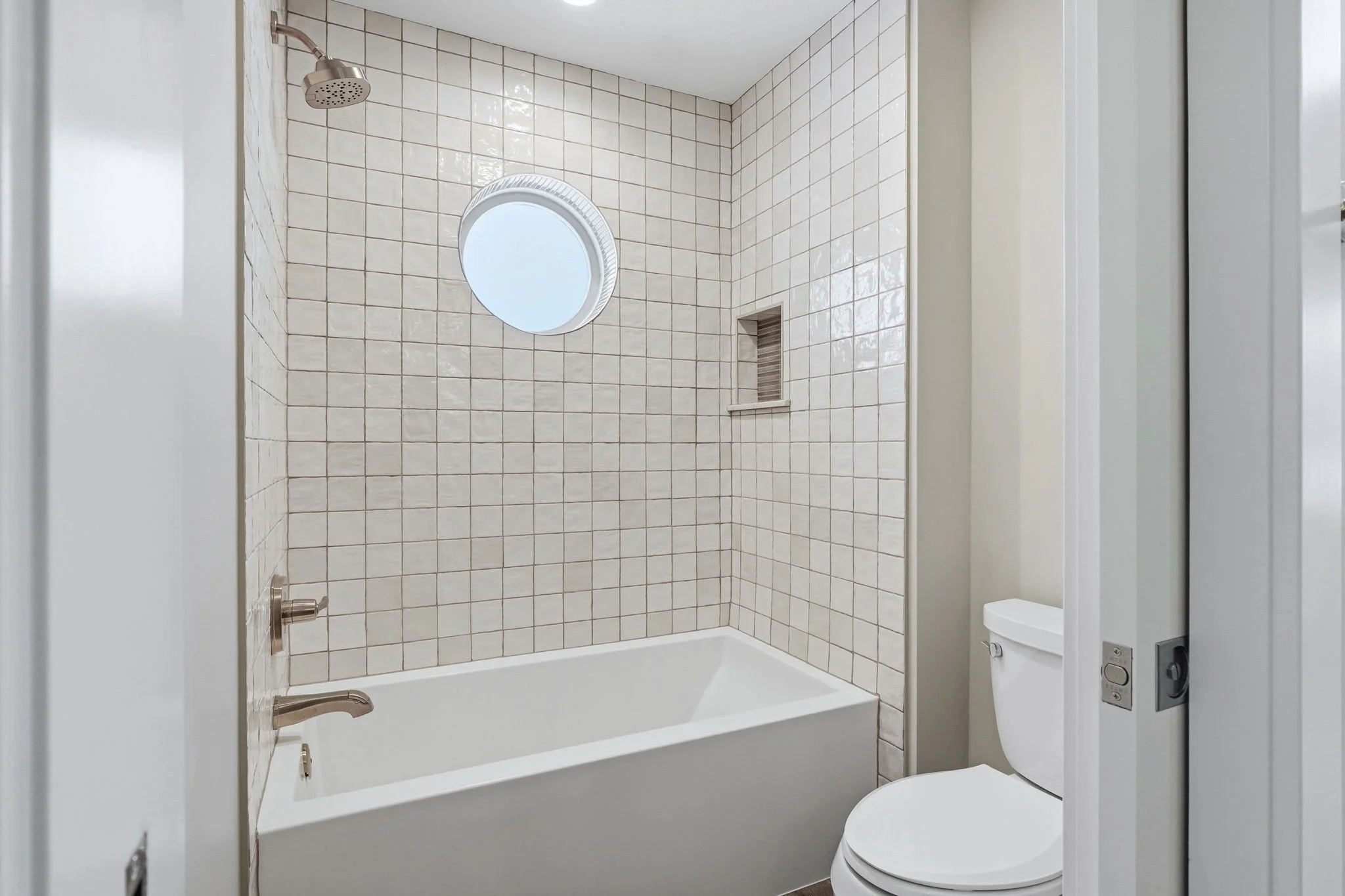 Bathroom with a white bathtub, tiled walls, a round window, a showerhead, and a toilet.
