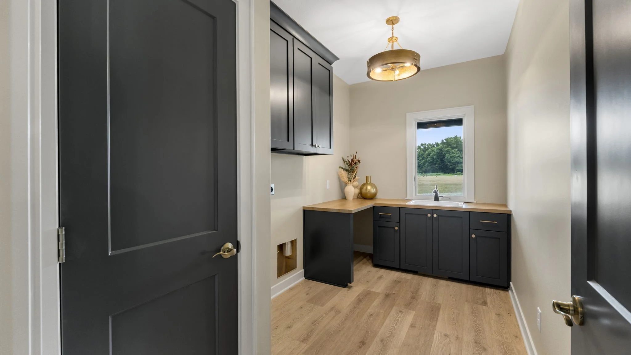 Empty laundry or utility room with black cabinets, beige walls, wood floor, small window showing green trees outside, decorative vases on the counter, ceiling light fixture.