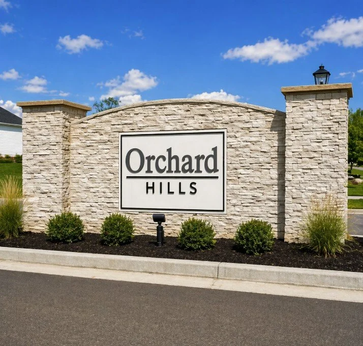 Entrance sign for Orchard Hills neighborhood, made of stone with black text on a white background, under a blue sky with some clouds.