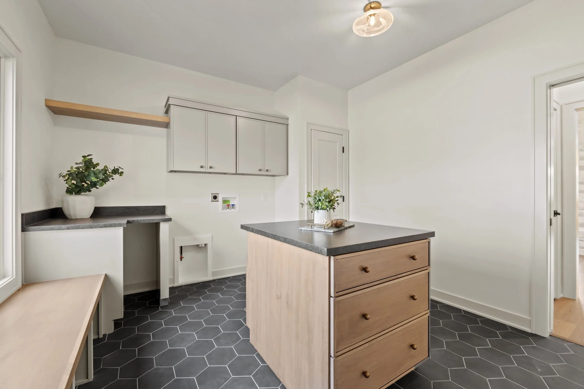 Empty kitchen with hexagon dark gray floor tiles, white walls, light wood and gray cabinets, an island with a dark countertop and a potted plant, and a small built-in table against the wall.