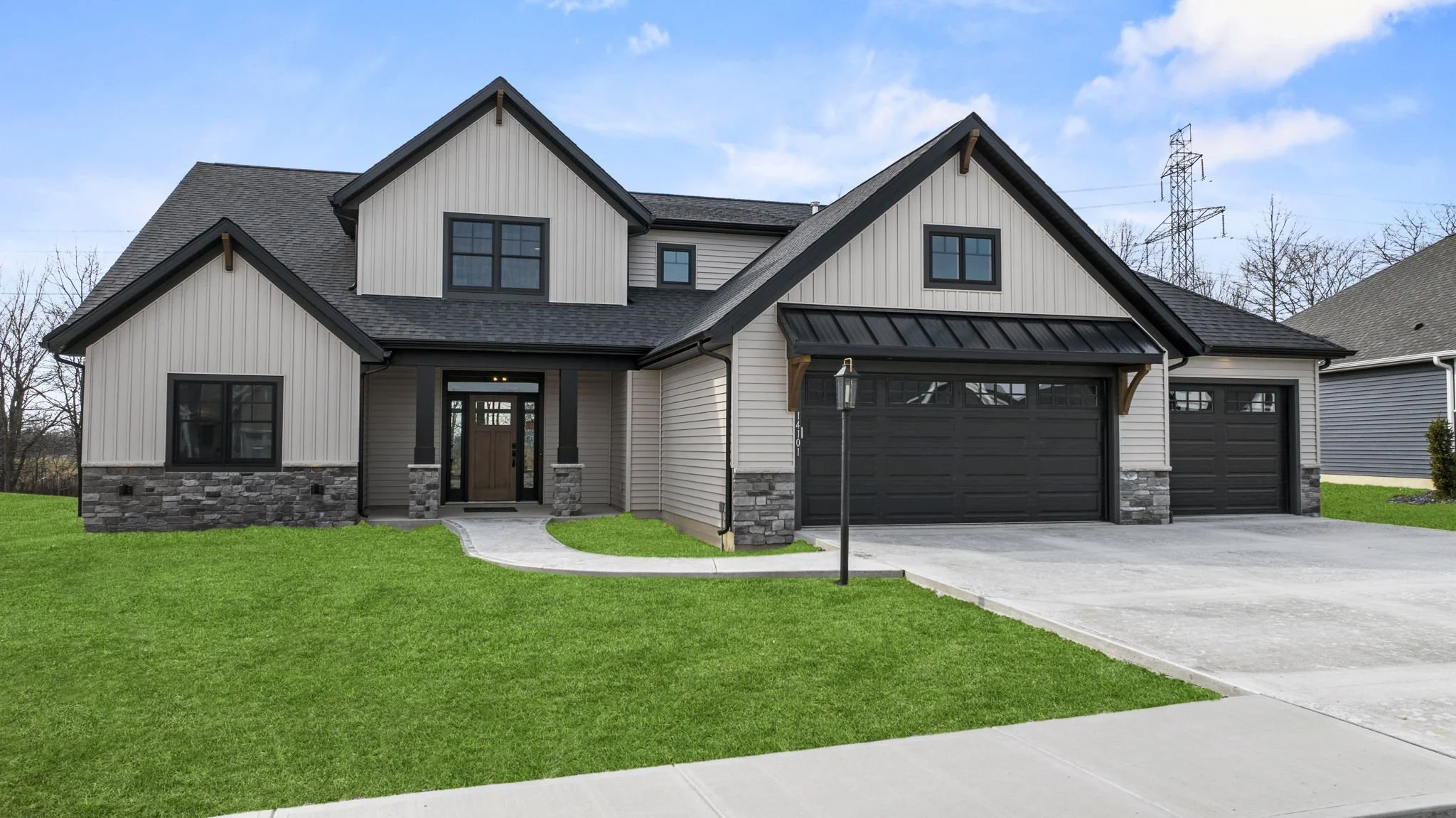 Front view of a modern house with beige siding, black window frames, a dark gray two-car garage door, stone accents, a wooden front door, and a well-maintained green lawn under a partly cloudy sky.