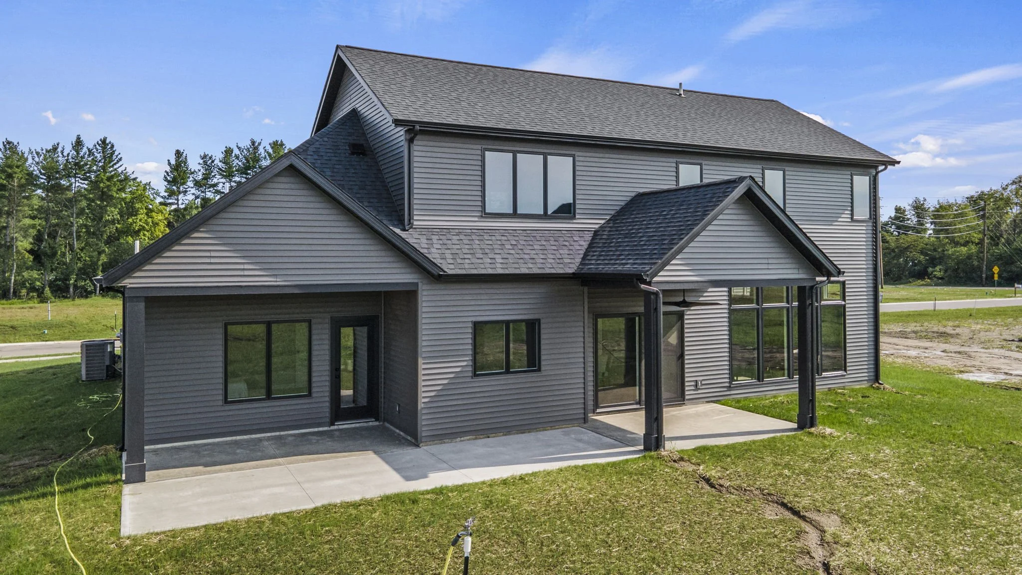 Newly built two-story house with gray siding and black trim, featuring a small porch and large windows, on a grassy lot with a concrete patio, under a blue sky with some clouds.