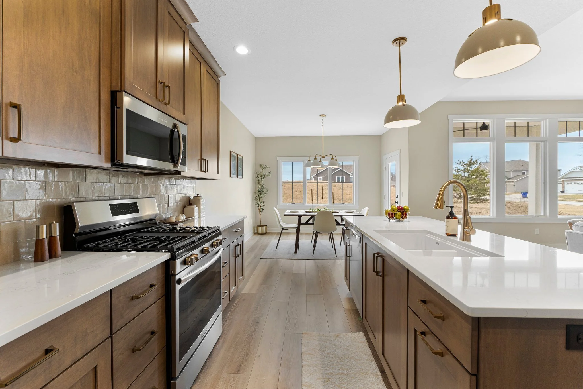 Modern kitchen with wooden cabinets, stainless steel appliances including a microwave and oven, white countertops, pendant and ceiling lights, and a dining area with large windows showing a residential neighborhood outside.
