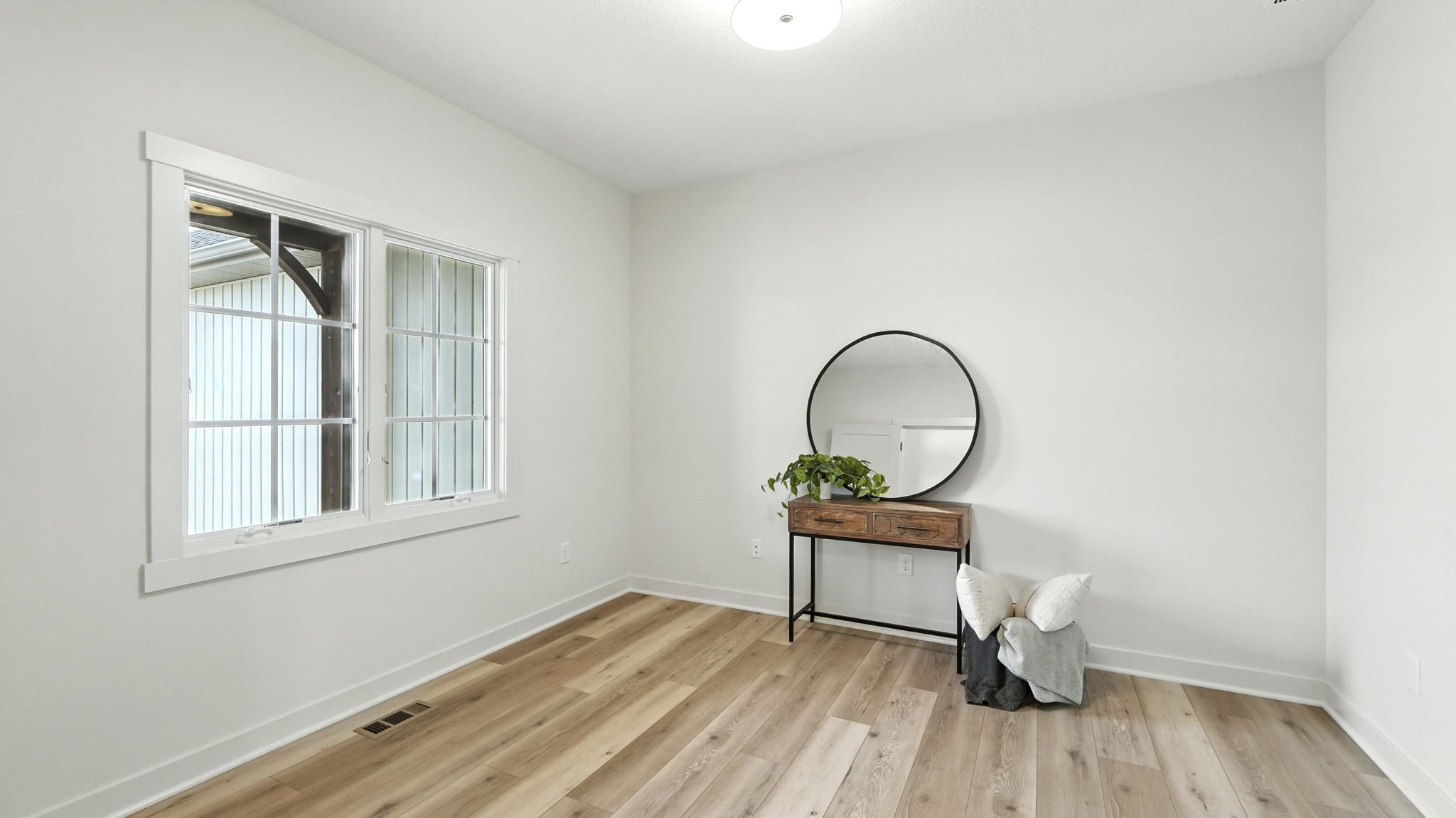 Empty room with white walls, wooden floors, a window, a small table with a plant and mirror, and folded pillows and blankets on the floor.