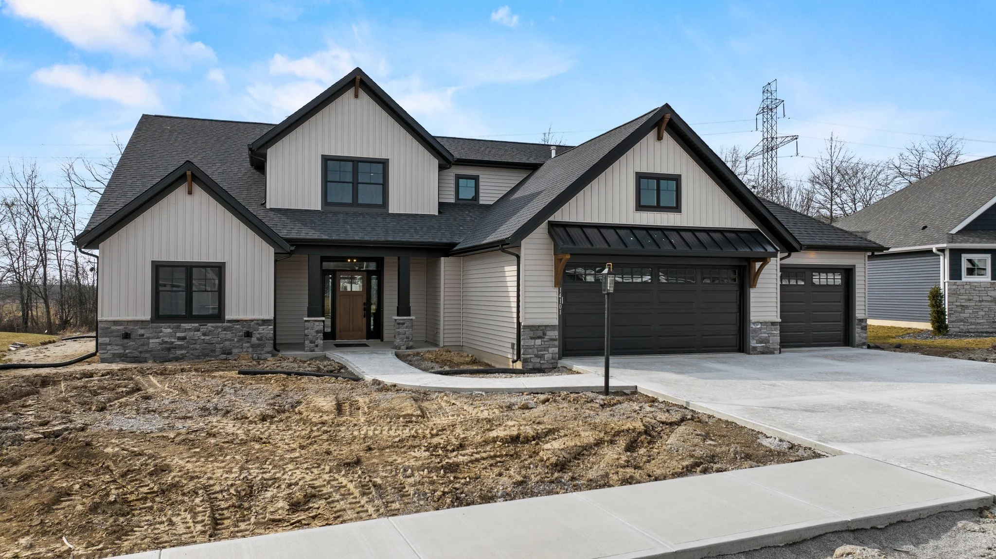 A modern two-story house with gray siding, black window frames, and a black garage door under a partly cloudy sky. The front yard has a concrete driveway and a small walkway, with some bare ground and construction debris in the foreground.